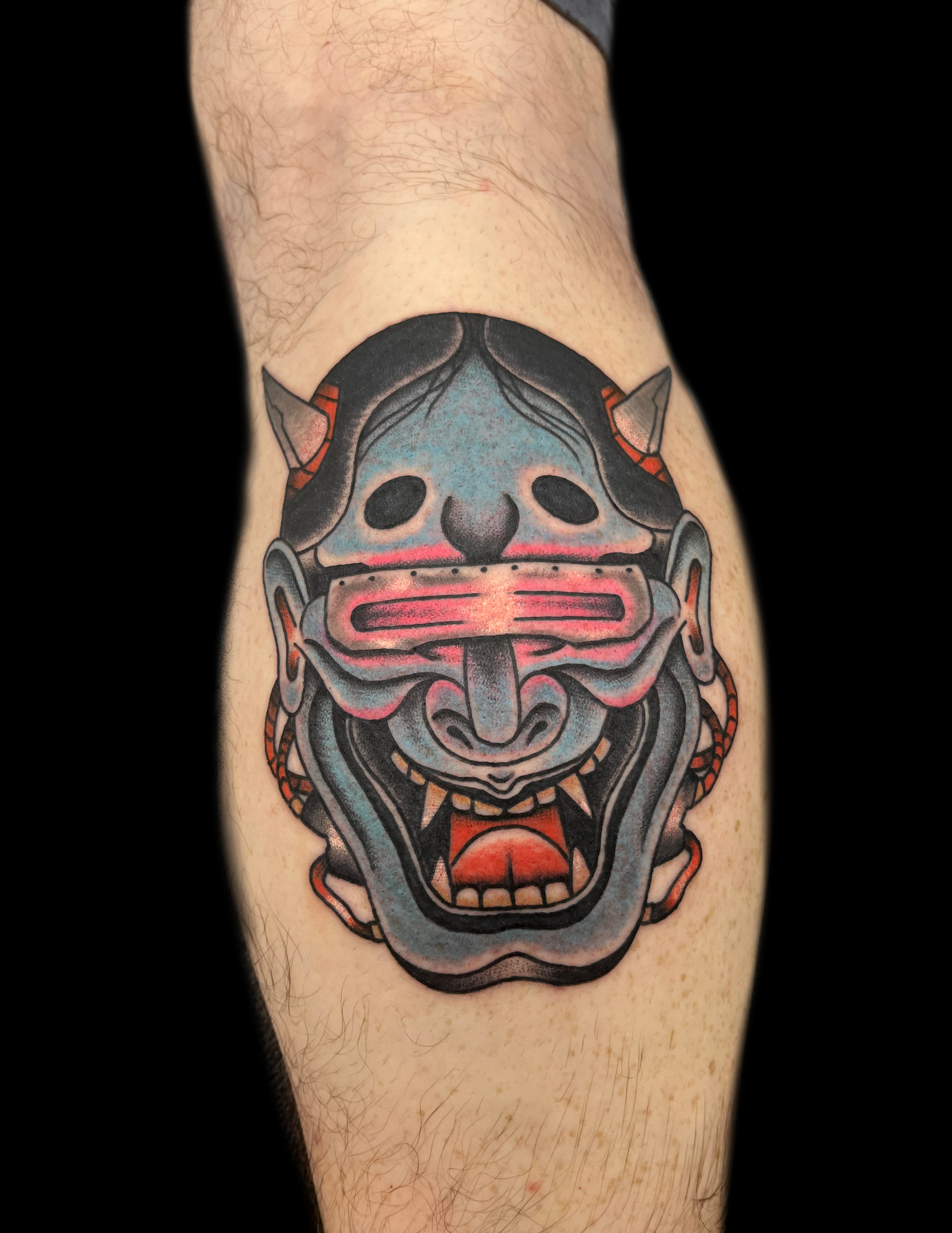 Tattoo of a blue cybernetic Oni mask on a leg. Pink visor, red mouth, small horns.