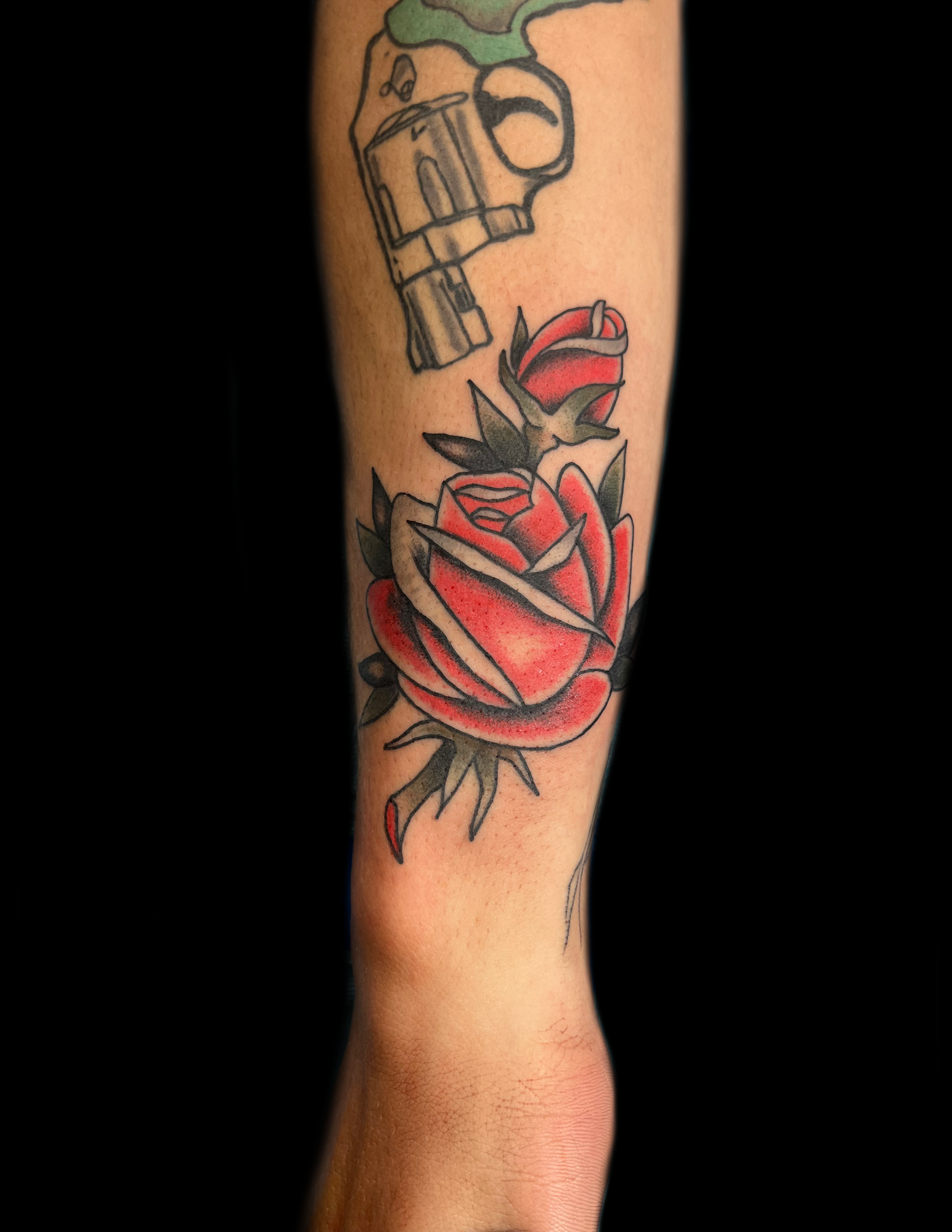 Tattoo of a red rose and a gun on an arm. The rose has green leaves and is against a black background.