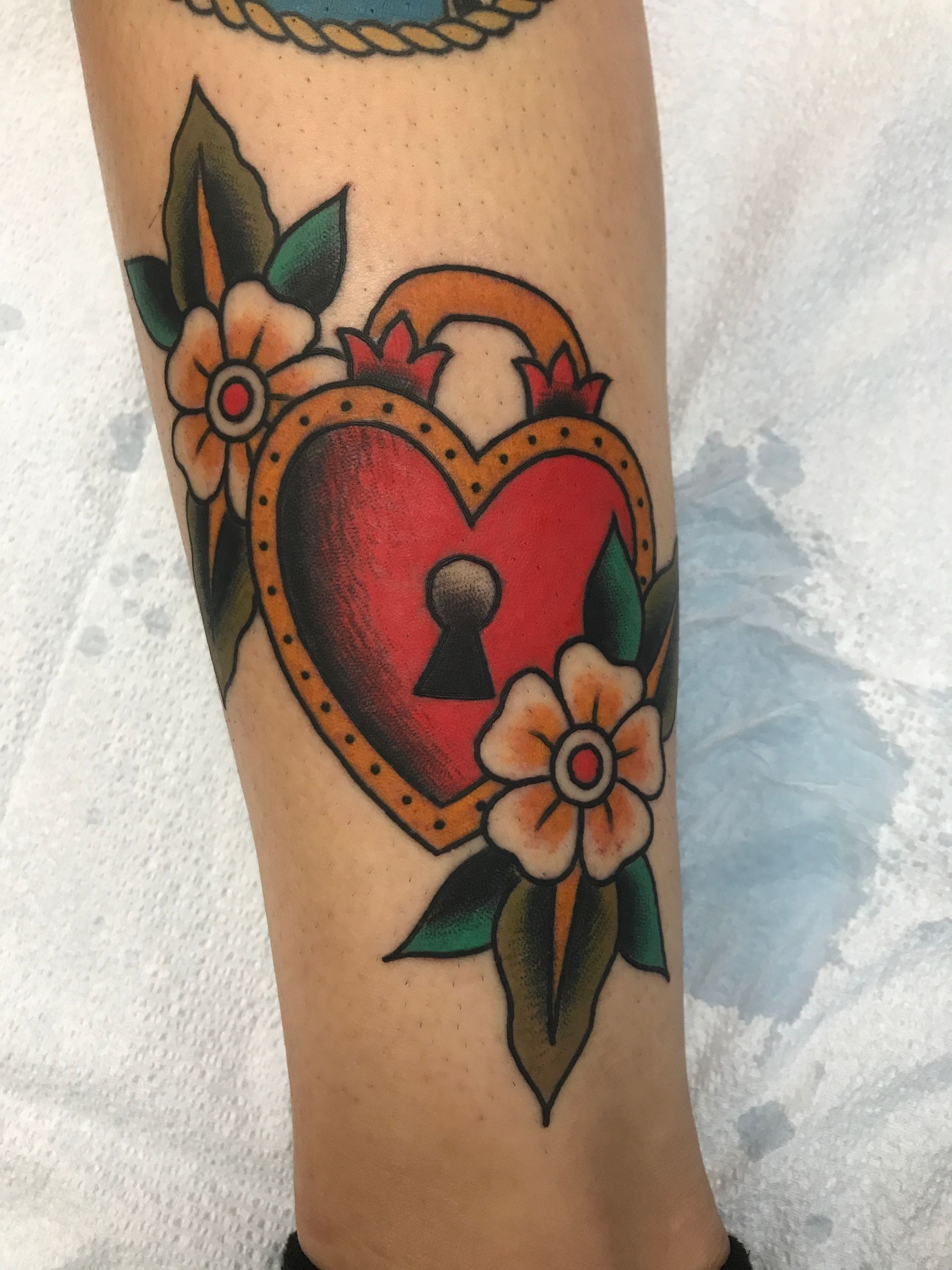 Heart-shaped lock tattoo in red and orange, surrounded by flowers and green leaves on a person's leg.