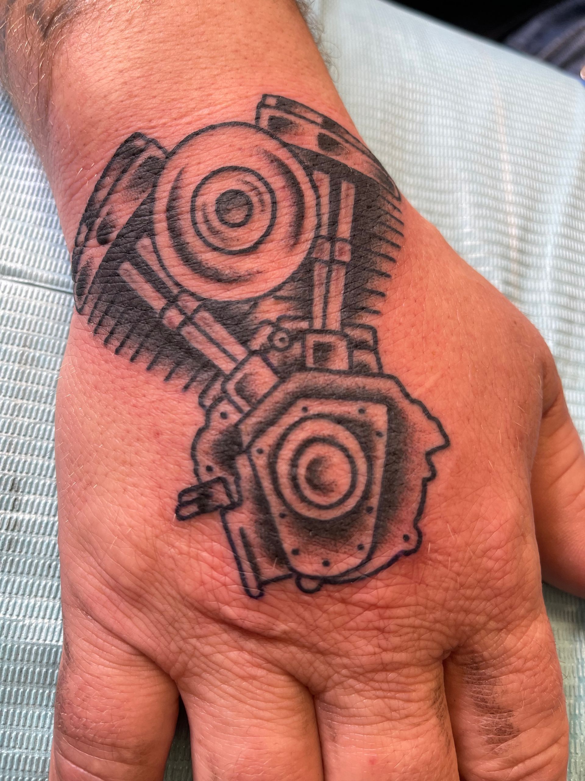 Tattoo of a motorcycle engine on the back of a hand, in black ink.