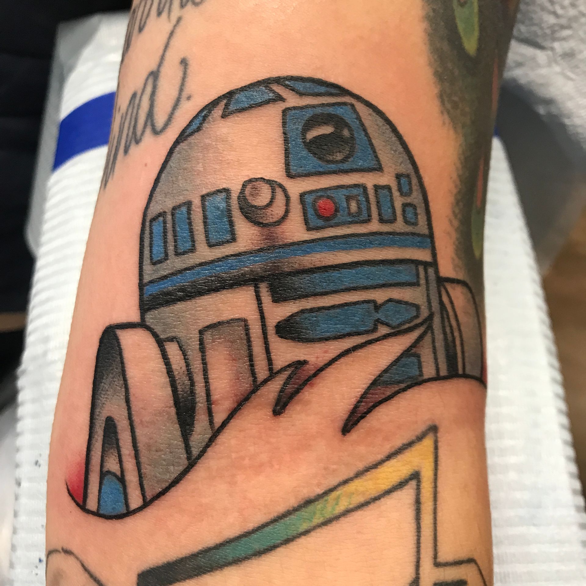 R2-D2 tattoo on an arm, with blue, gray, and black details, a colorful design below.