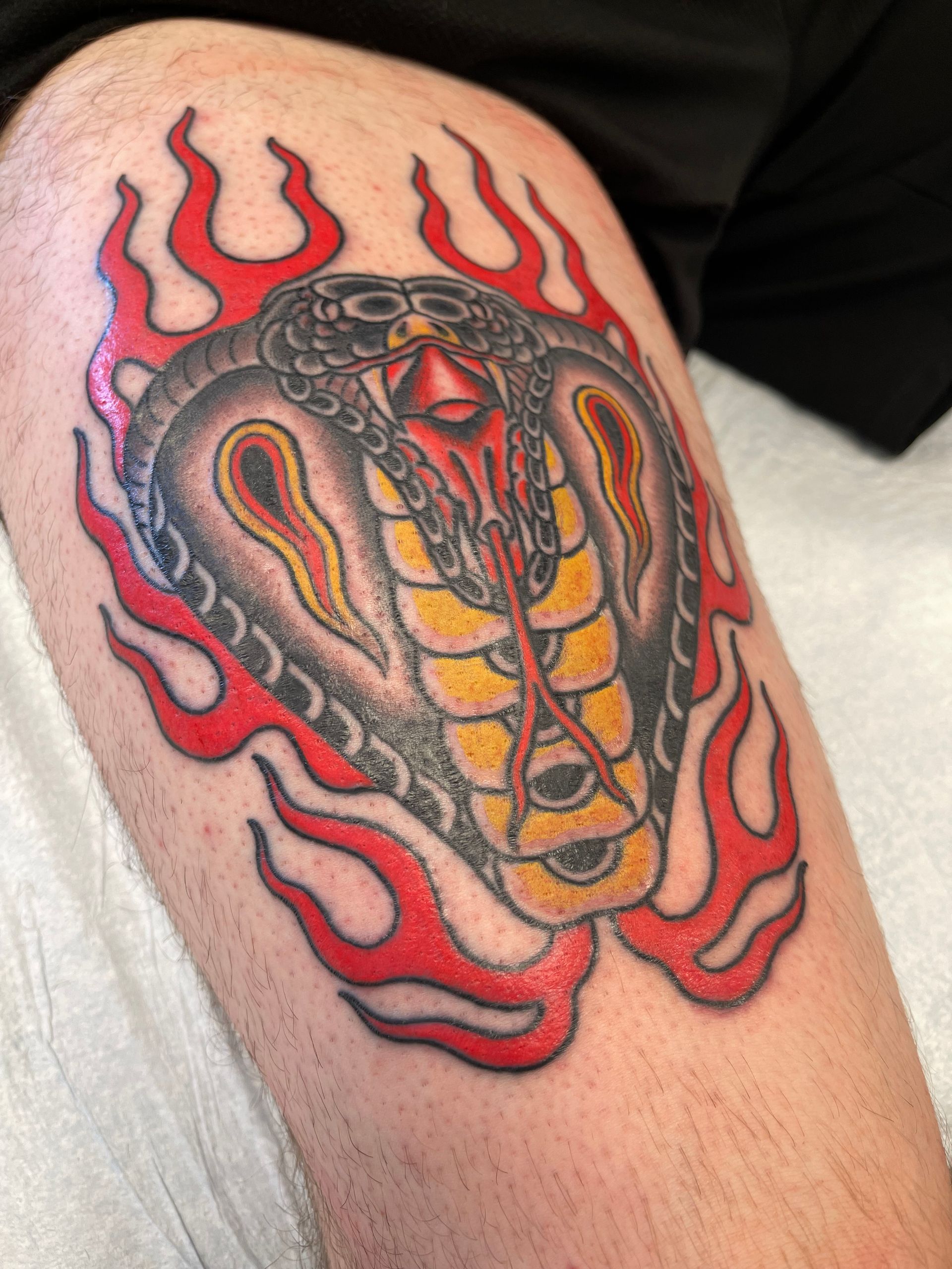 Tattoo of a cobra head surrounded by red flames on a leg. The cobra is in shades of grey, yellow, and red.