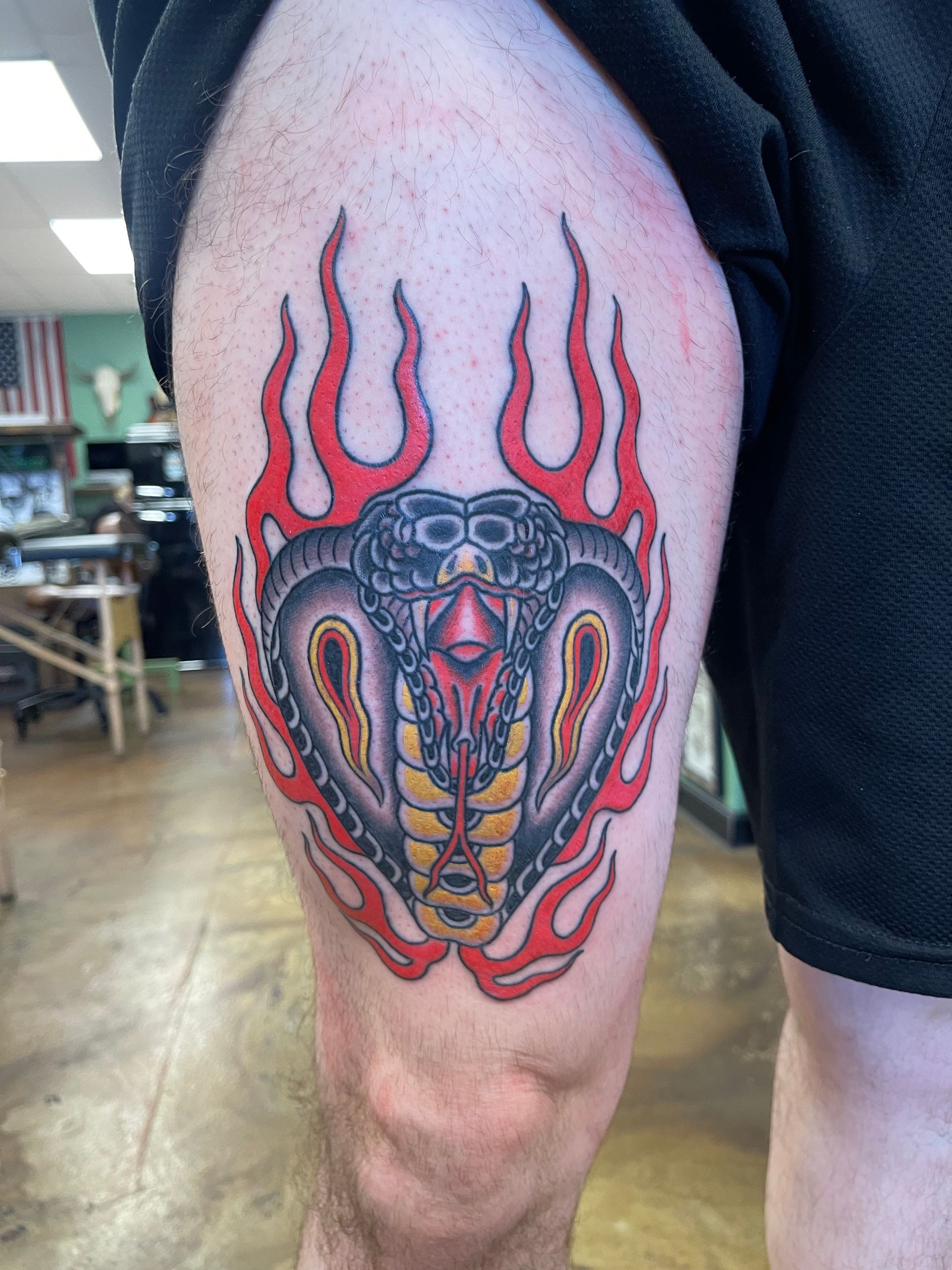Cobra tattoo on a thigh, with flames. Red, black, yellow, and gray colors. In a tattoo parlor setting.