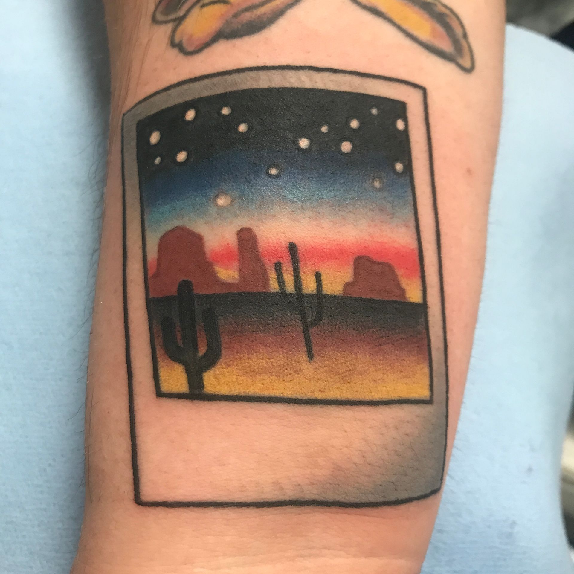 Tattoo of a desert sunset in a Polaroid frame, with cacti and mountains under a starry sky; on a person's arm.