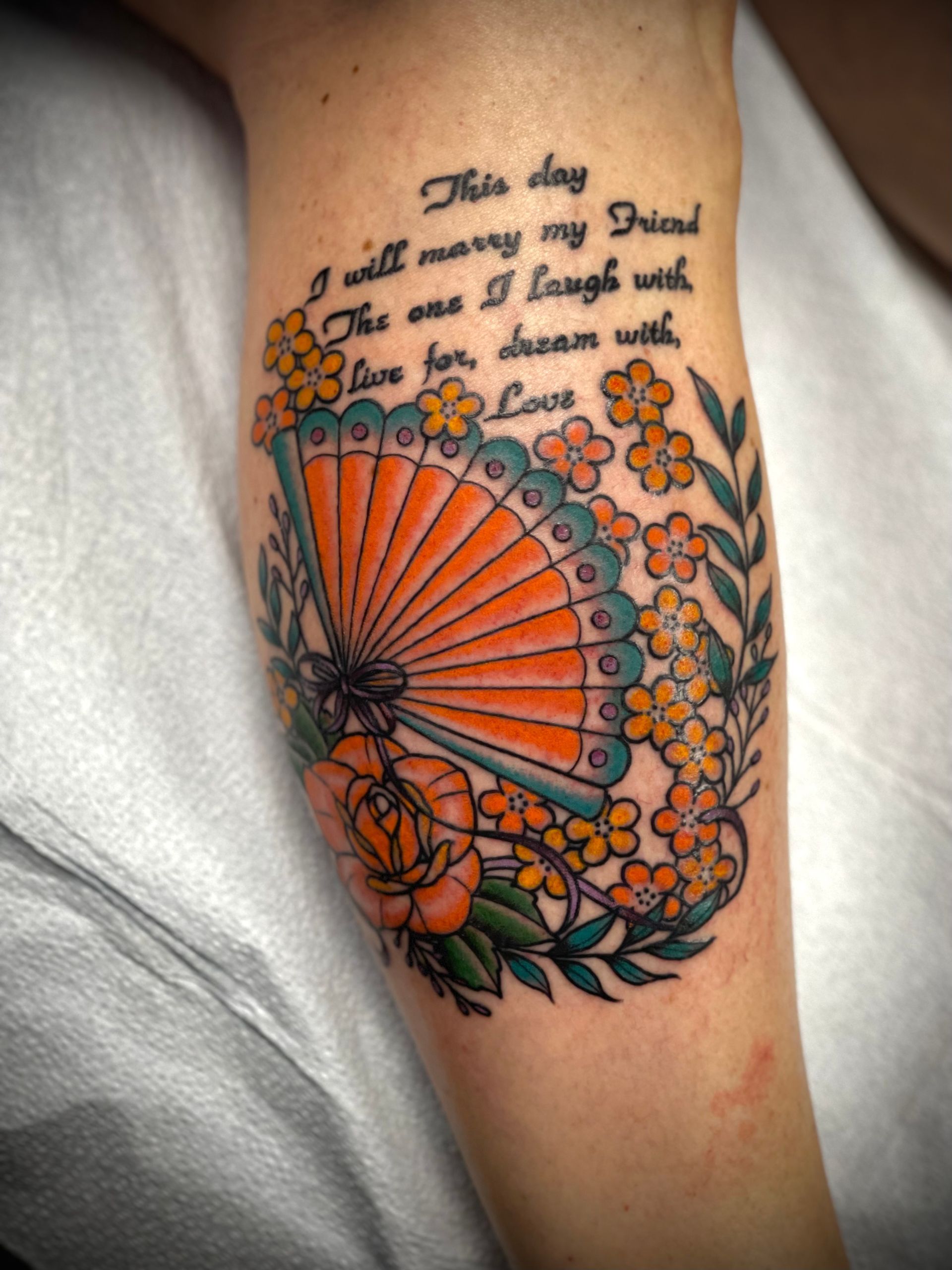 Tattoo of a fan, flowers, and text on an arm: 