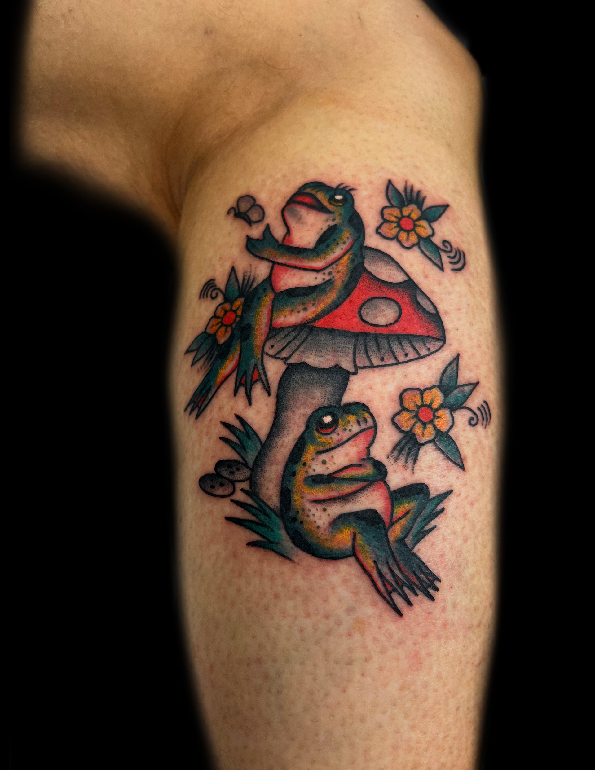 Tattoo of two frogs near a mushroom and flowers, on a leg. One frog sits atop the mushroom, the other below.