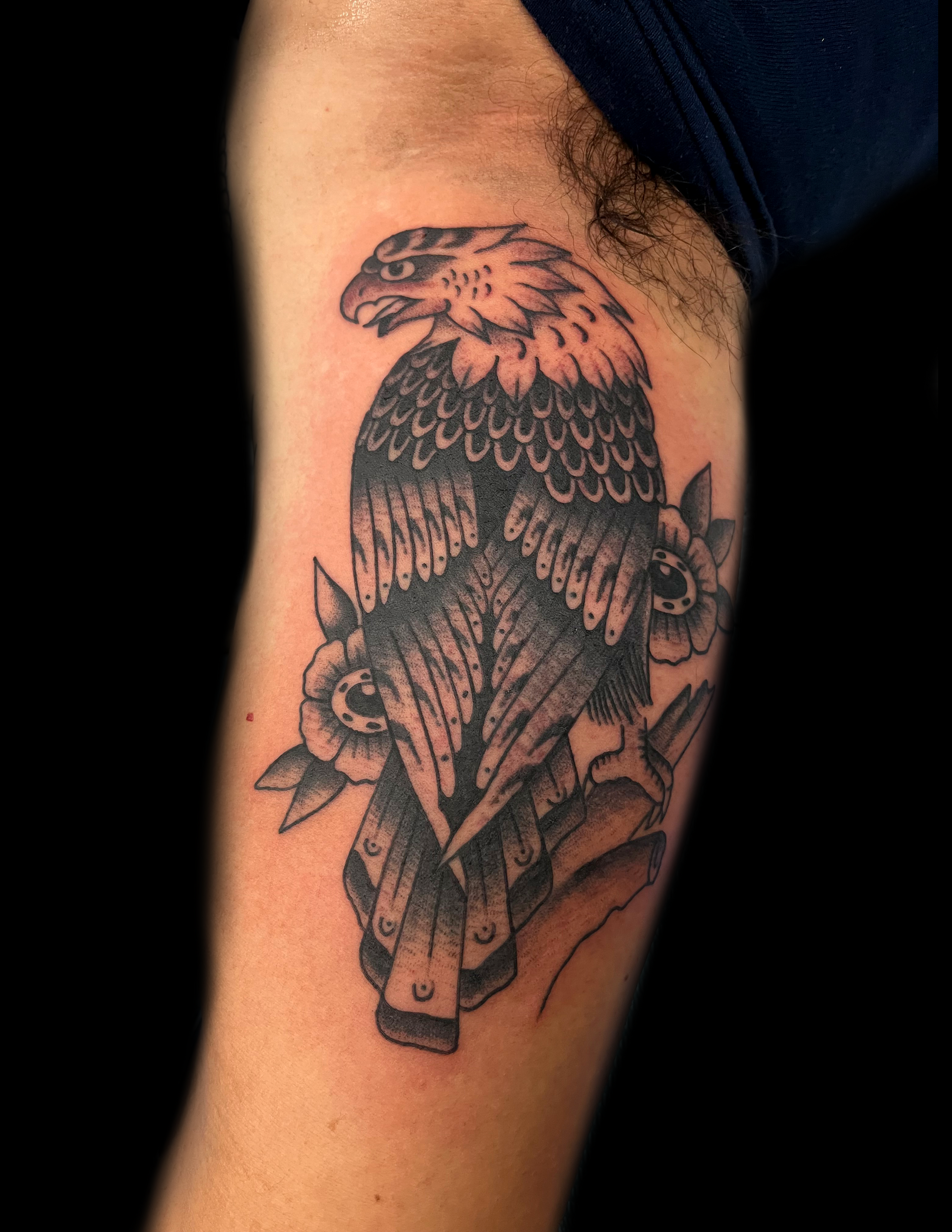 Black and white tattoo of an eagle perched on a branch, flowers on either side.