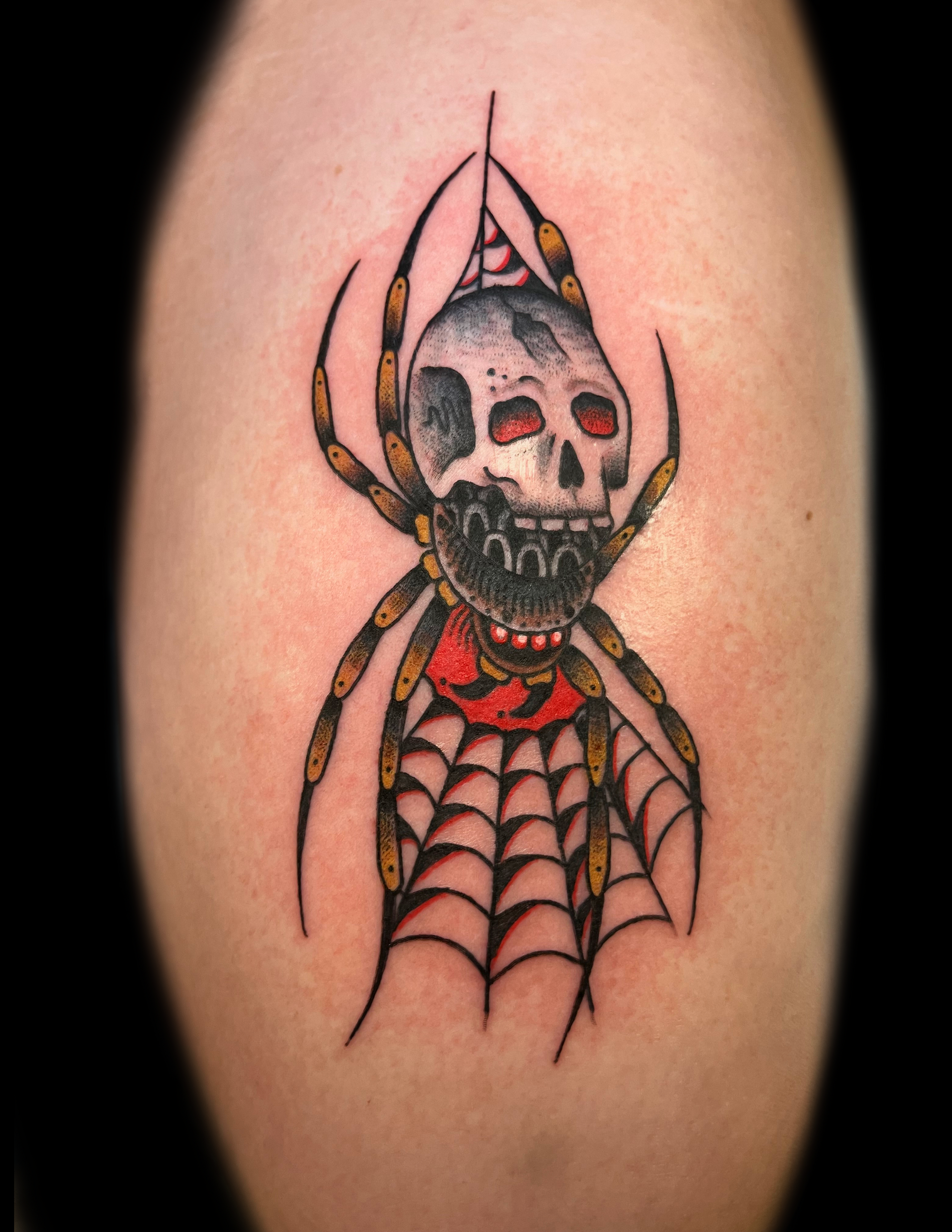 Tattoo of a skull-faced spider with a web on a person's arm. Red eyes, red collar, and black, gold, and white details.