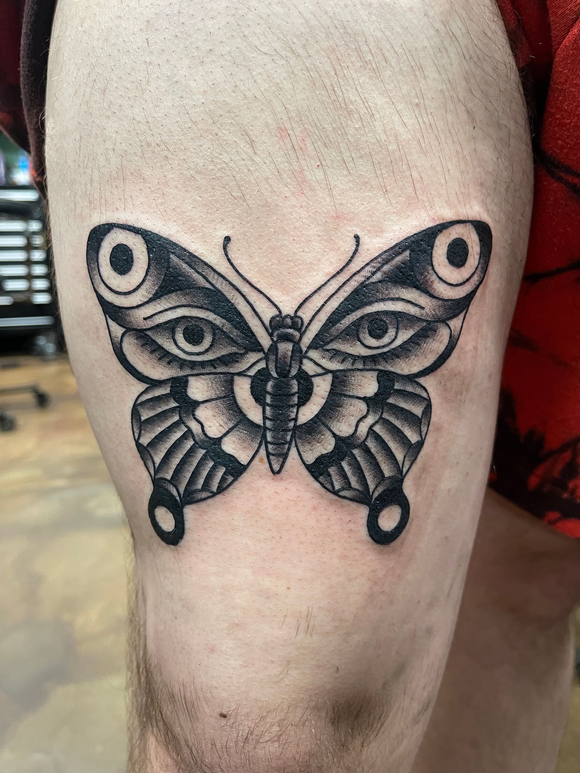 Tattoo of a black and gray butterfly with two eyes in the wings on a person's thigh.