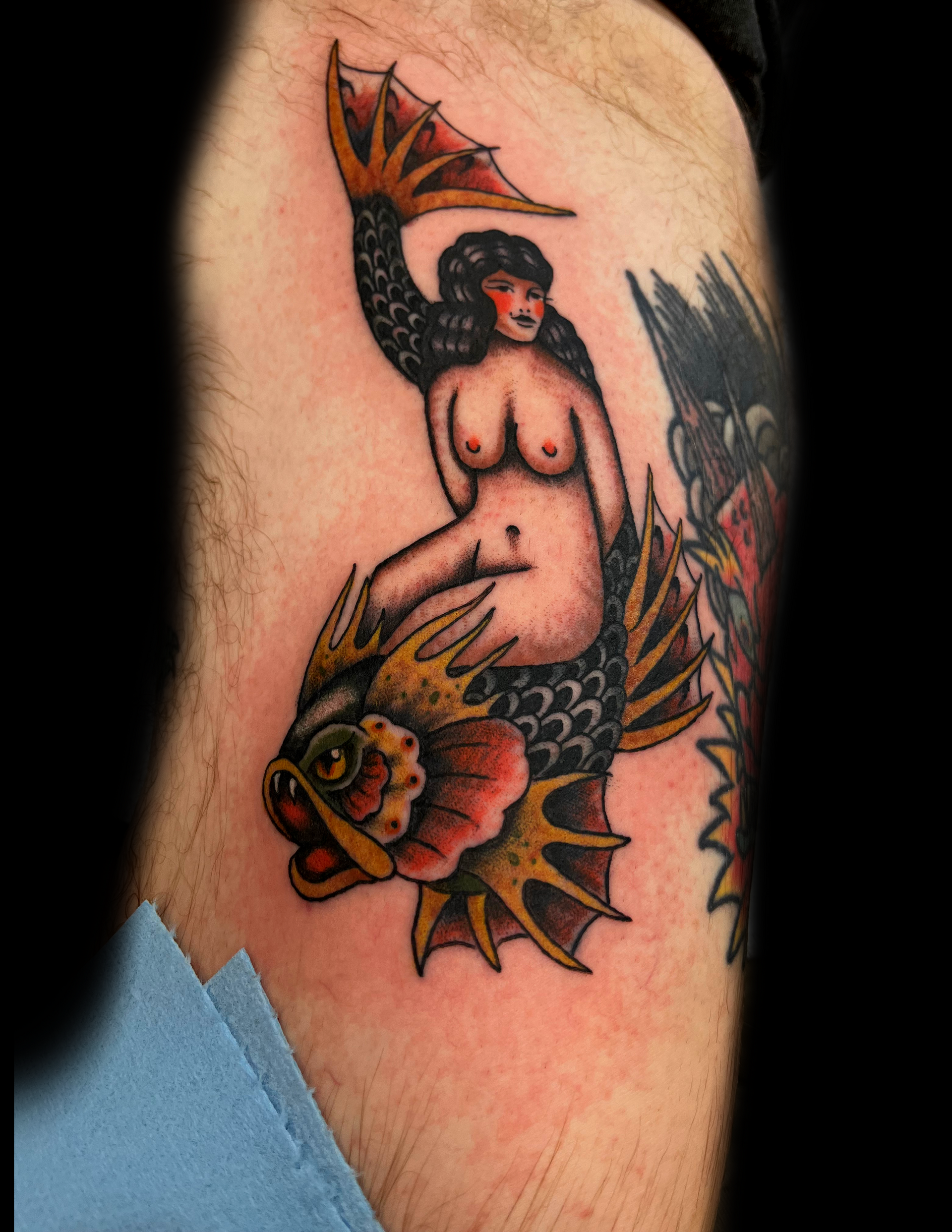 Naked mermaid on a fish tattoo in red, yellow, and black ink on skin.