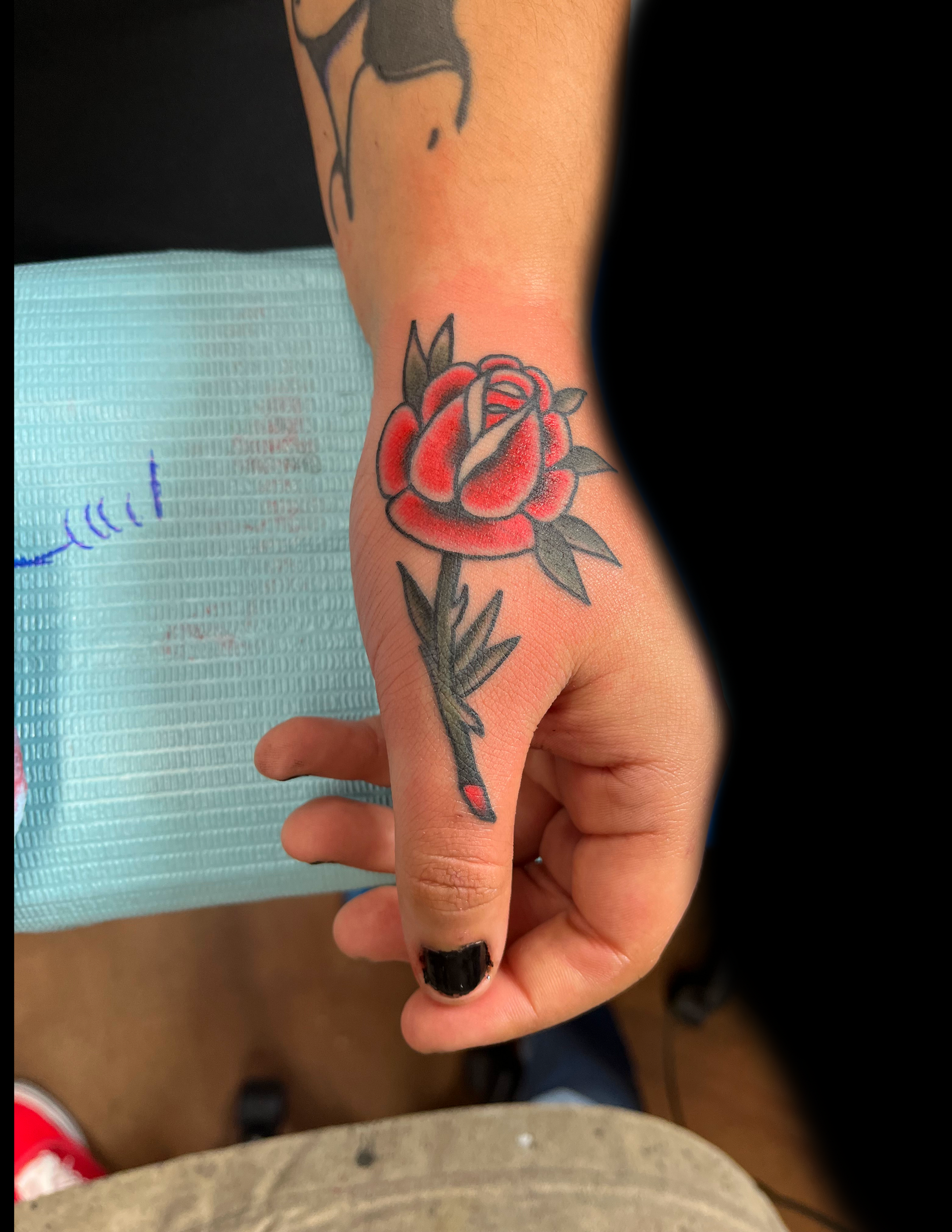 Hand with a red rose tattoo on the back, stem down the thumb, black nail polish.