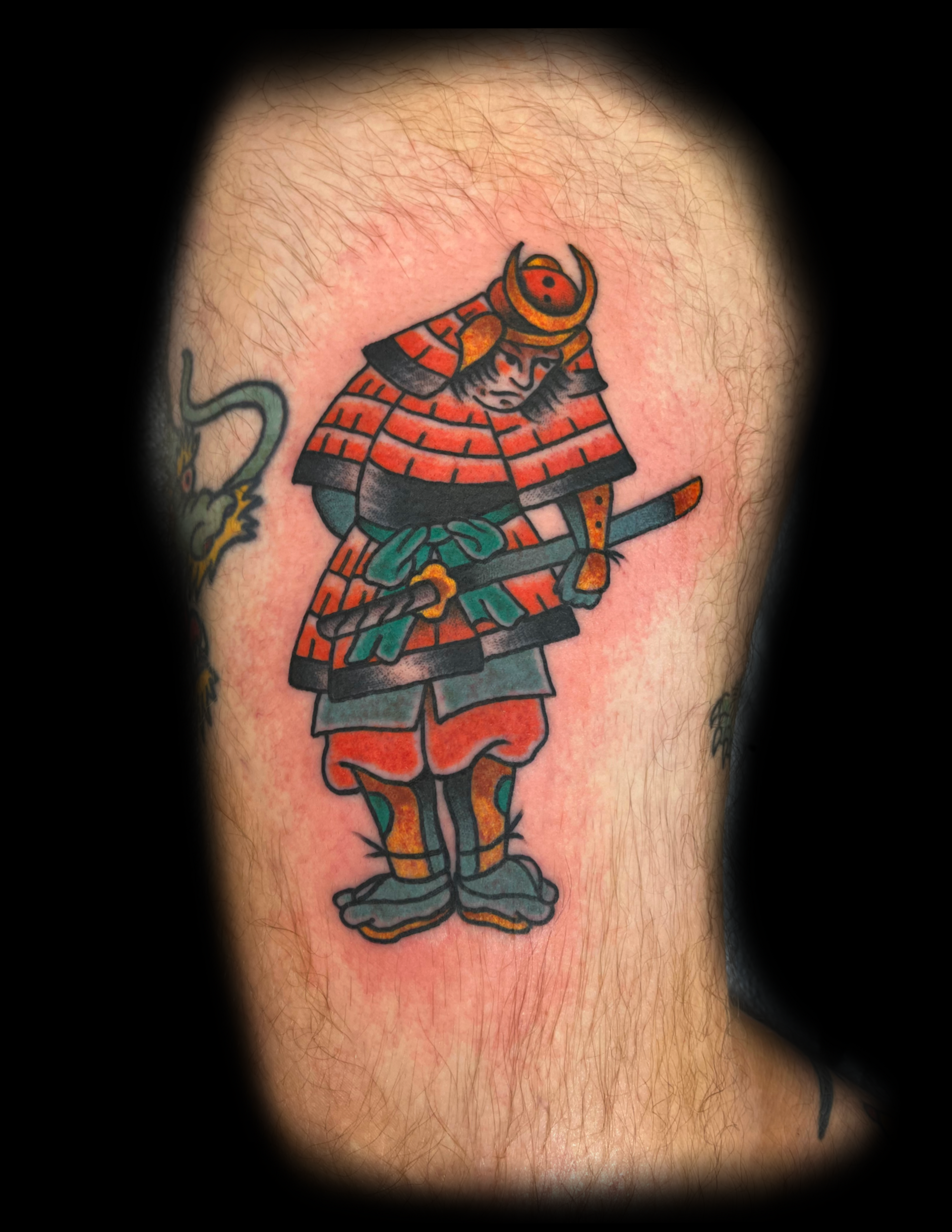 Tattoo of a traditional samurai warrior in red and black armor, holding a katana, on a person's arm.