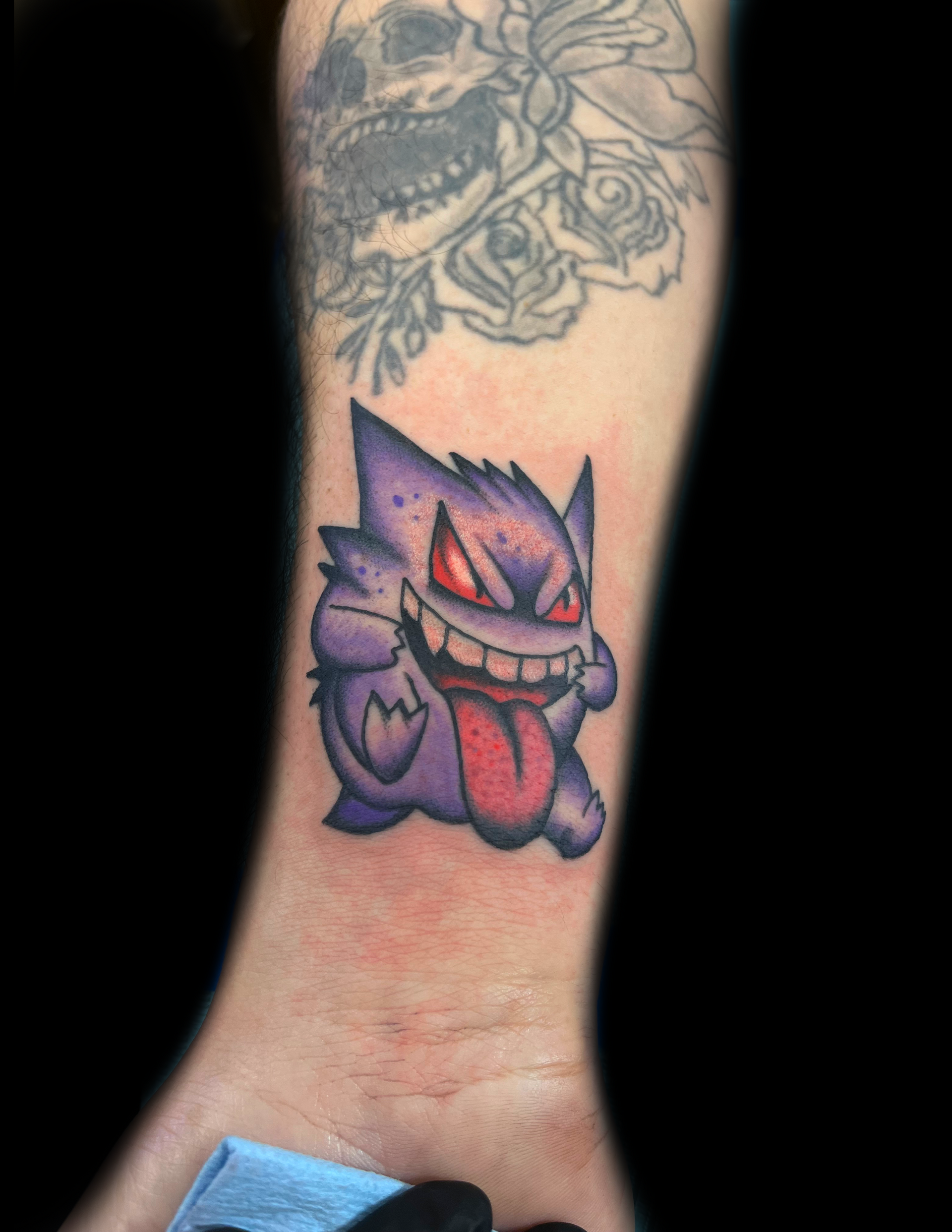 Gengar tattoo on wrist, purple and red, with a smiling, menacing expression. Another faded tattoo above.