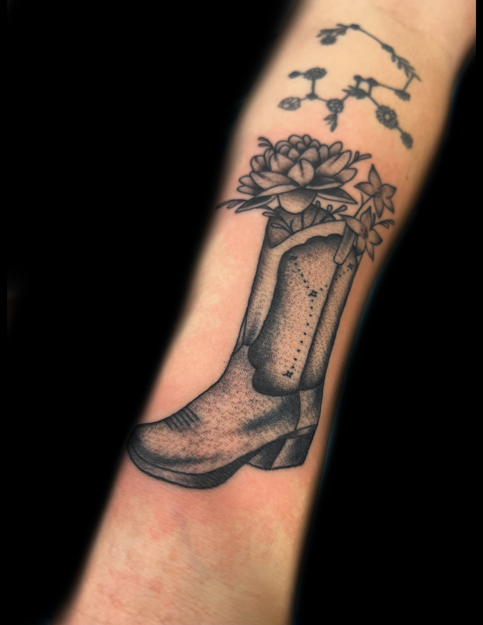 Black and white tattoo of a cowboy boot with flowers growing out, on a person's arm.