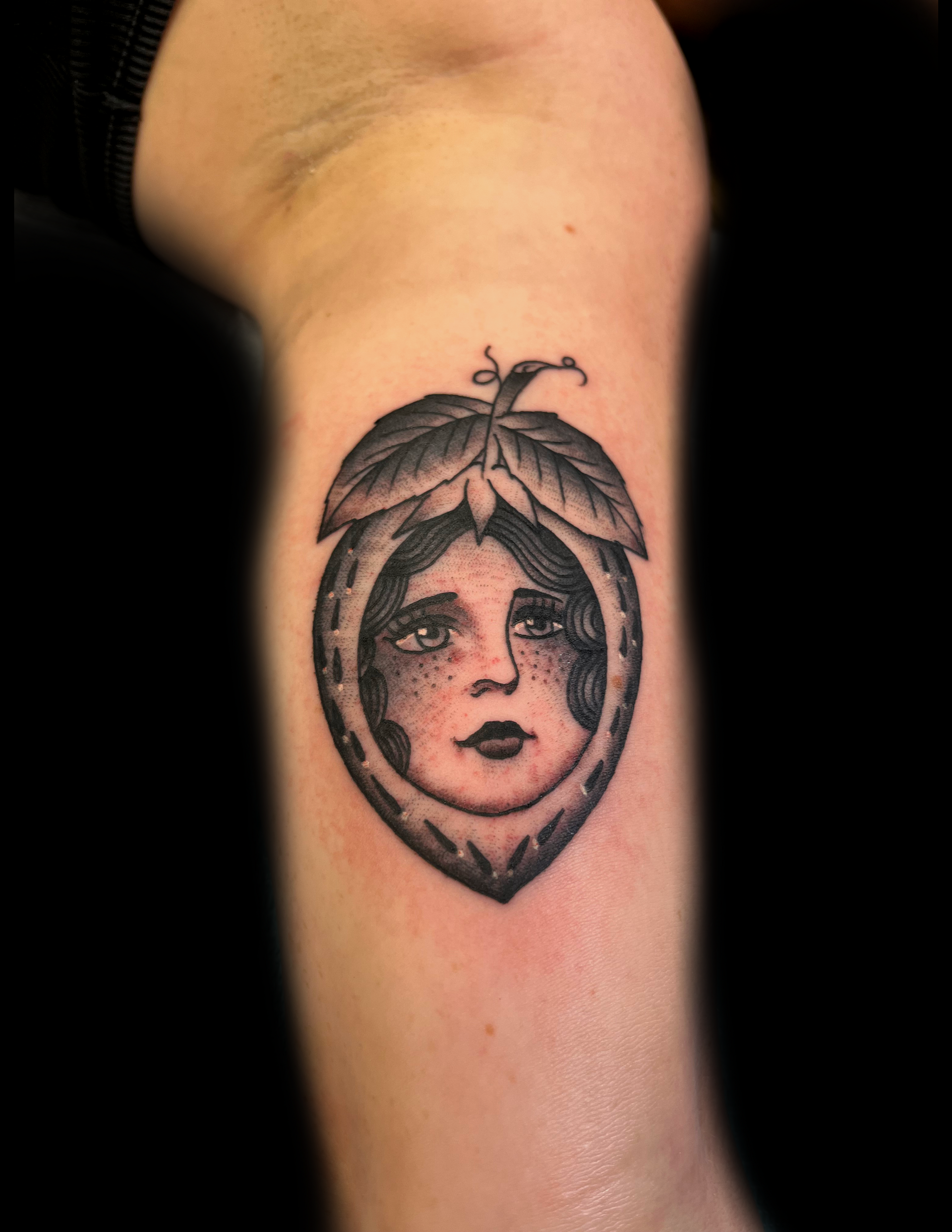 Tattoo of a face inside a stylized peach. Black and gray, on a wrist.