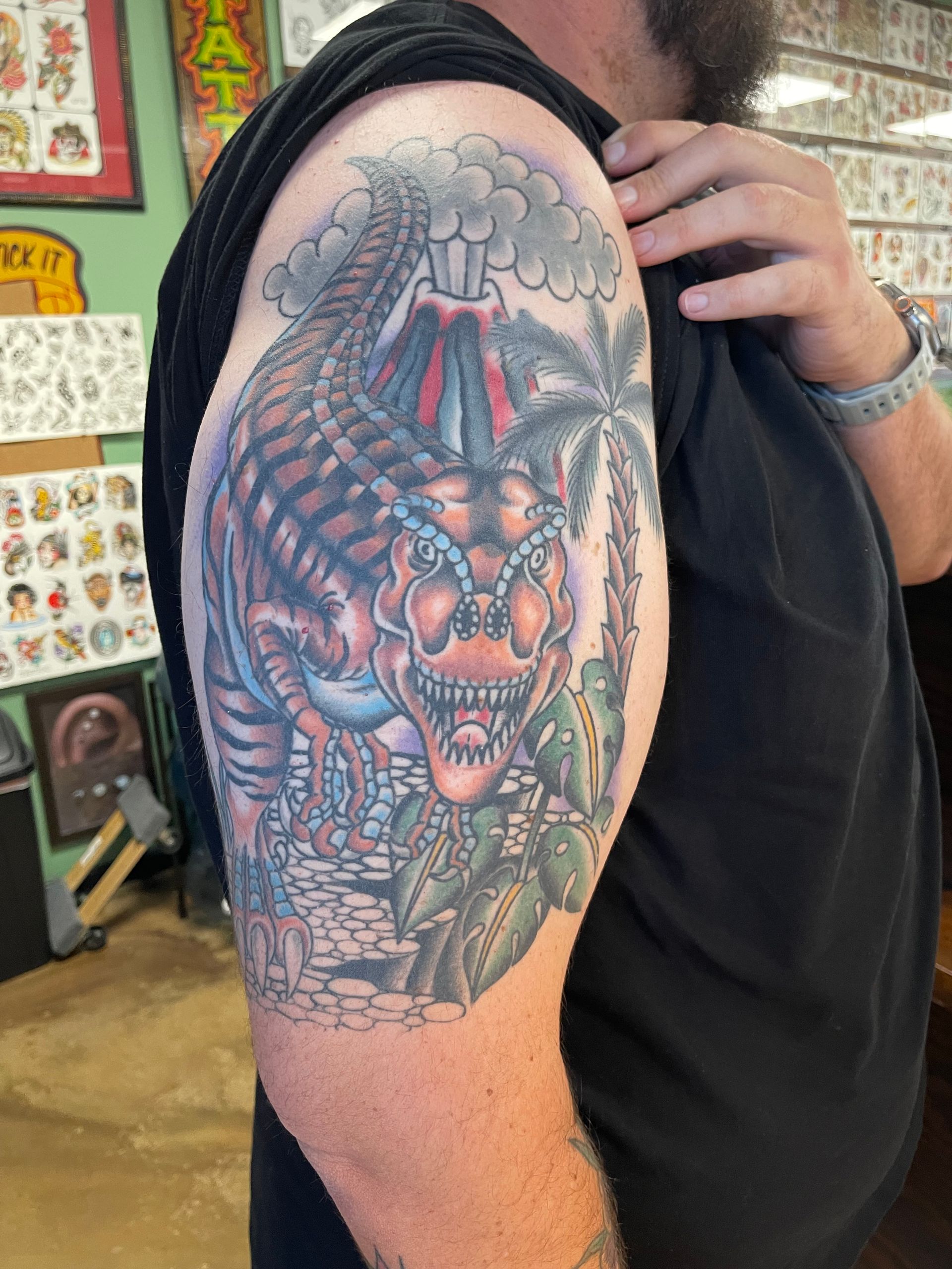 Tattoo of a Tyrannosaurus Rex by a volcano with palm tree and foliage on a man's arm.