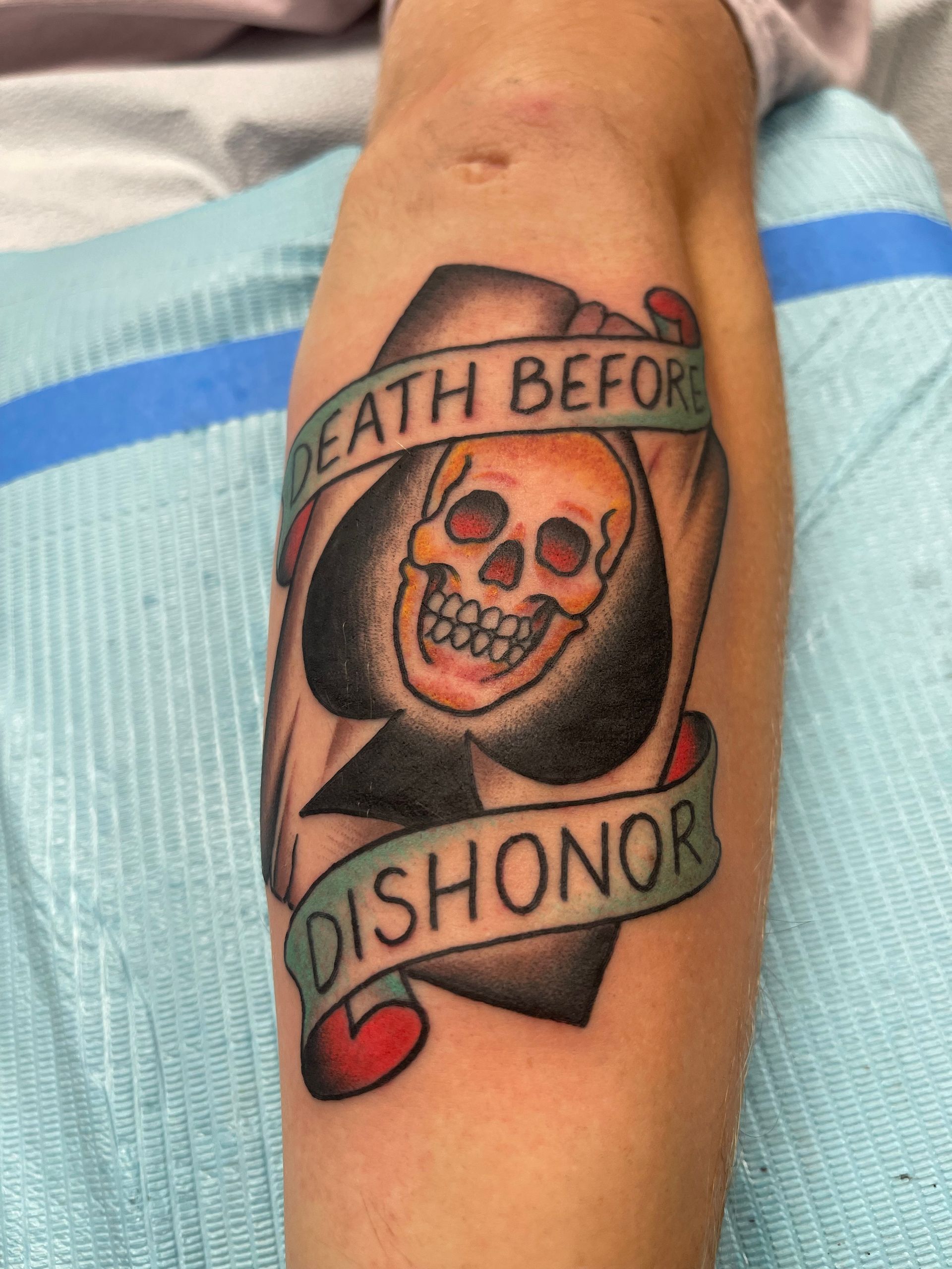Tattoo on forearm, a skull within a spade card, with banner reading 
