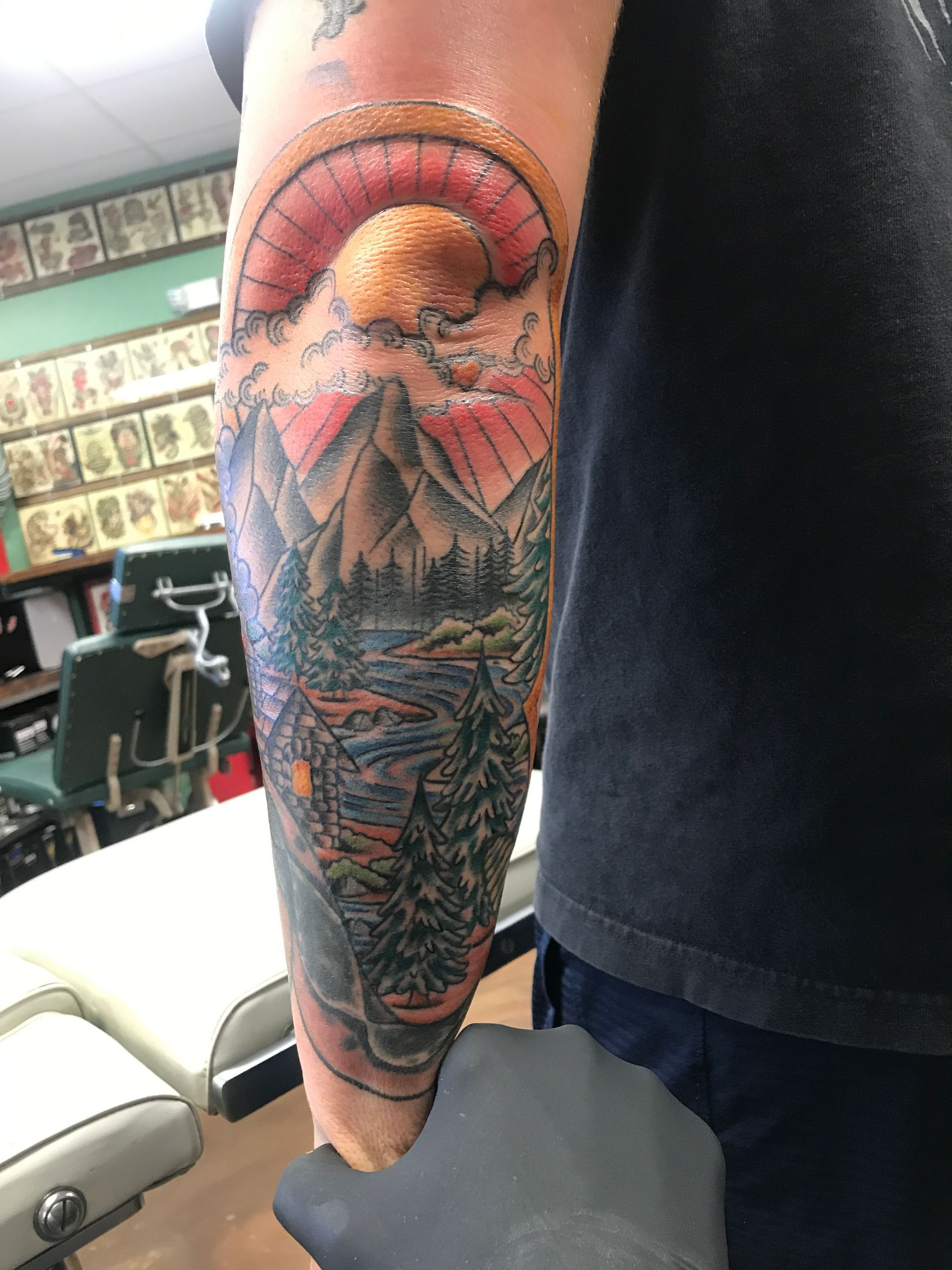 Tattoo of a mountain scene on a forearm. Features a red sunset, peaks, trees, and water.