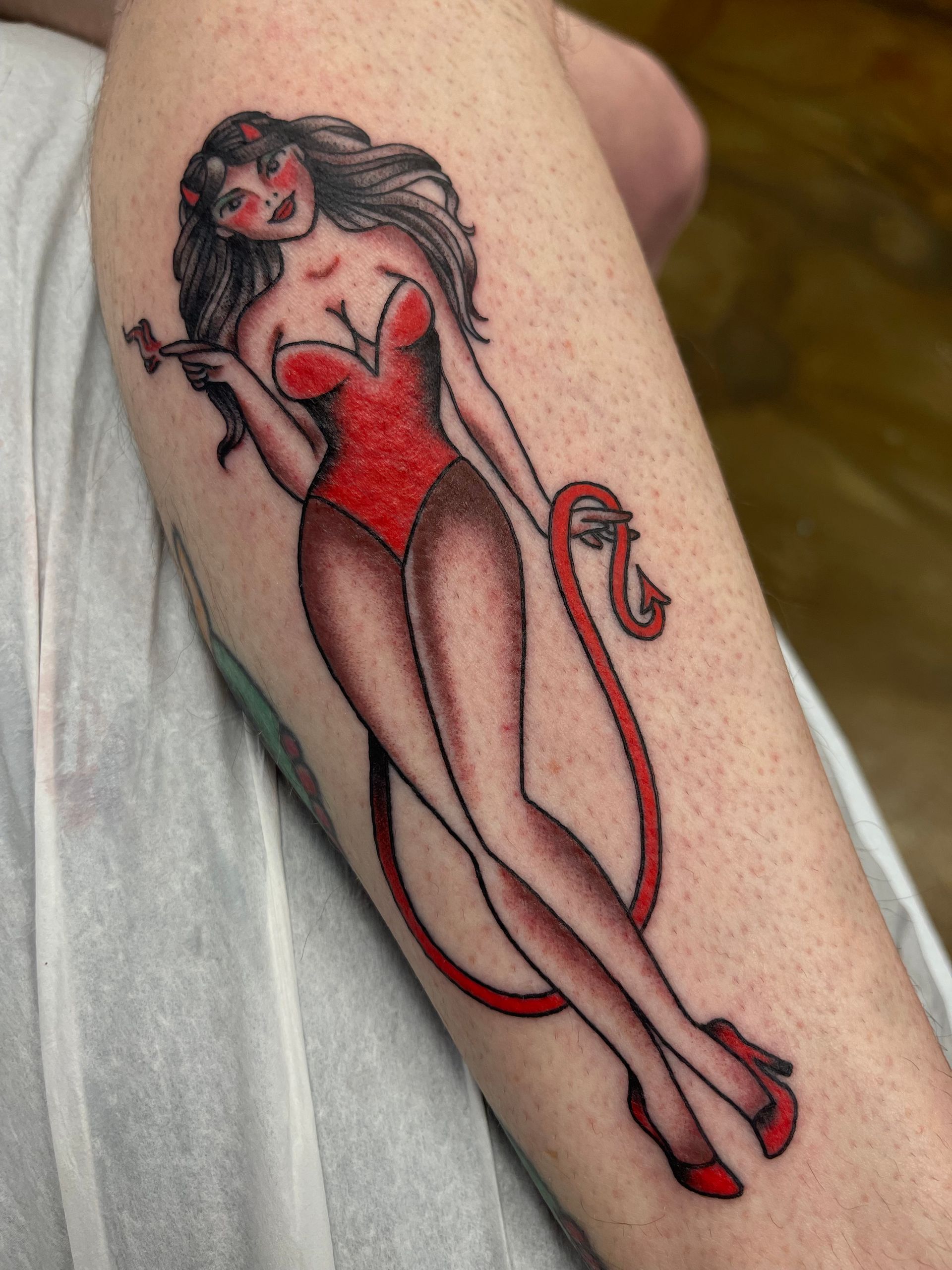 Tattoo of a woman in a red bodysuit, holding a flower, with a tail, and horns on leg.