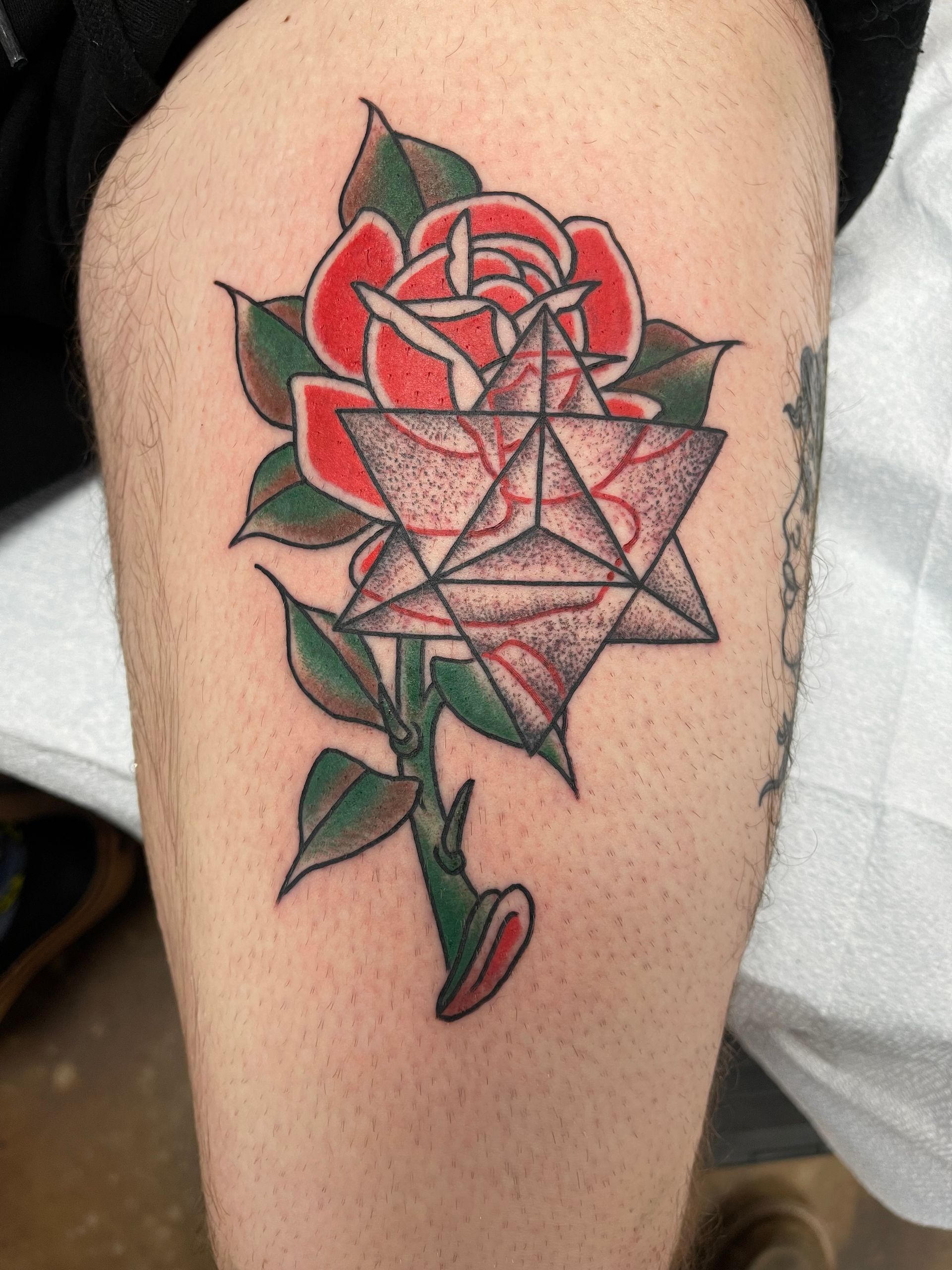 Tattoo on a leg: a red rose with a geometric star design in front; green leaves and stem.