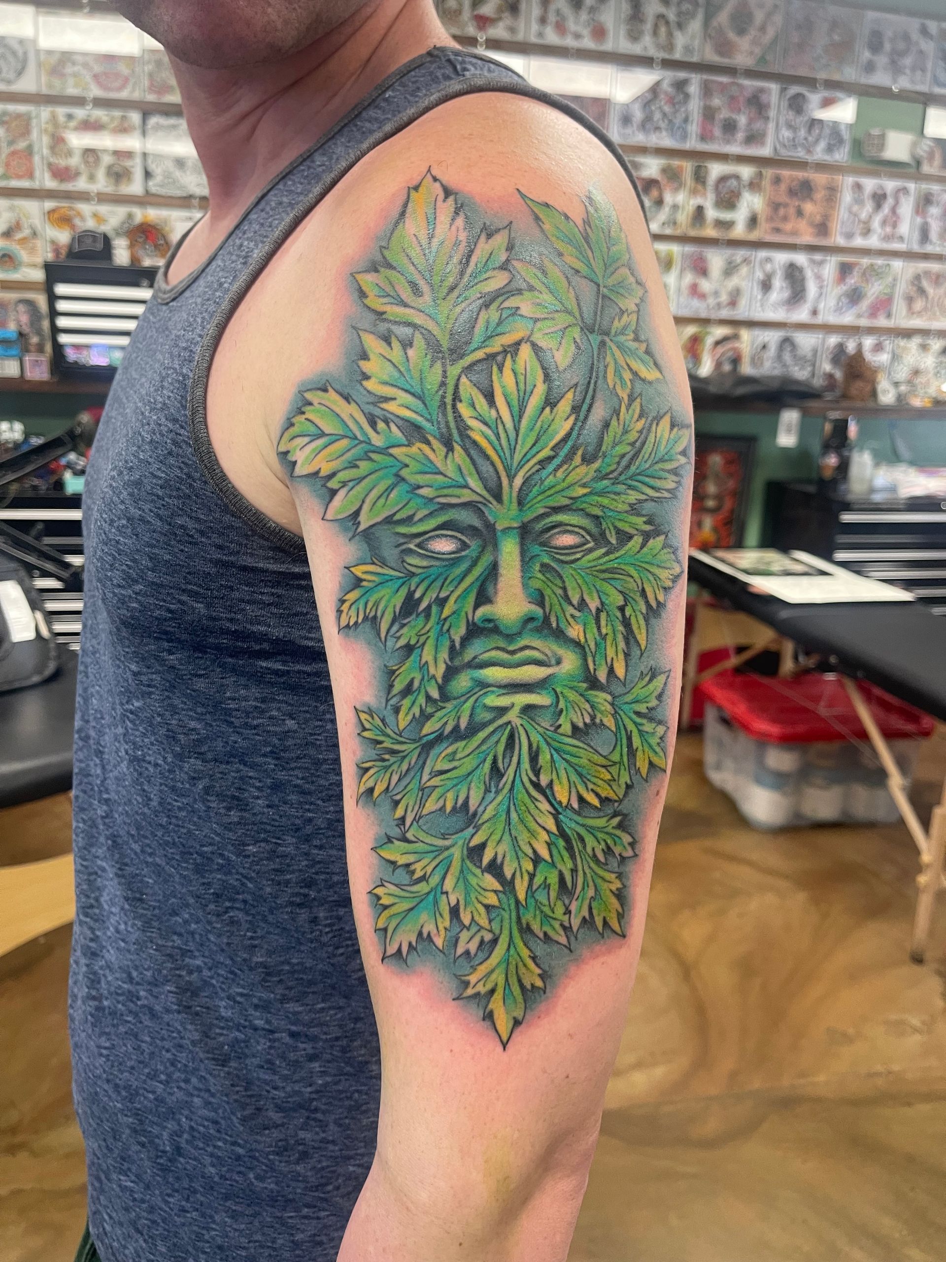 Tattoo of a green face made of leaves on a person's upper arm, inside a tattoo parlor.