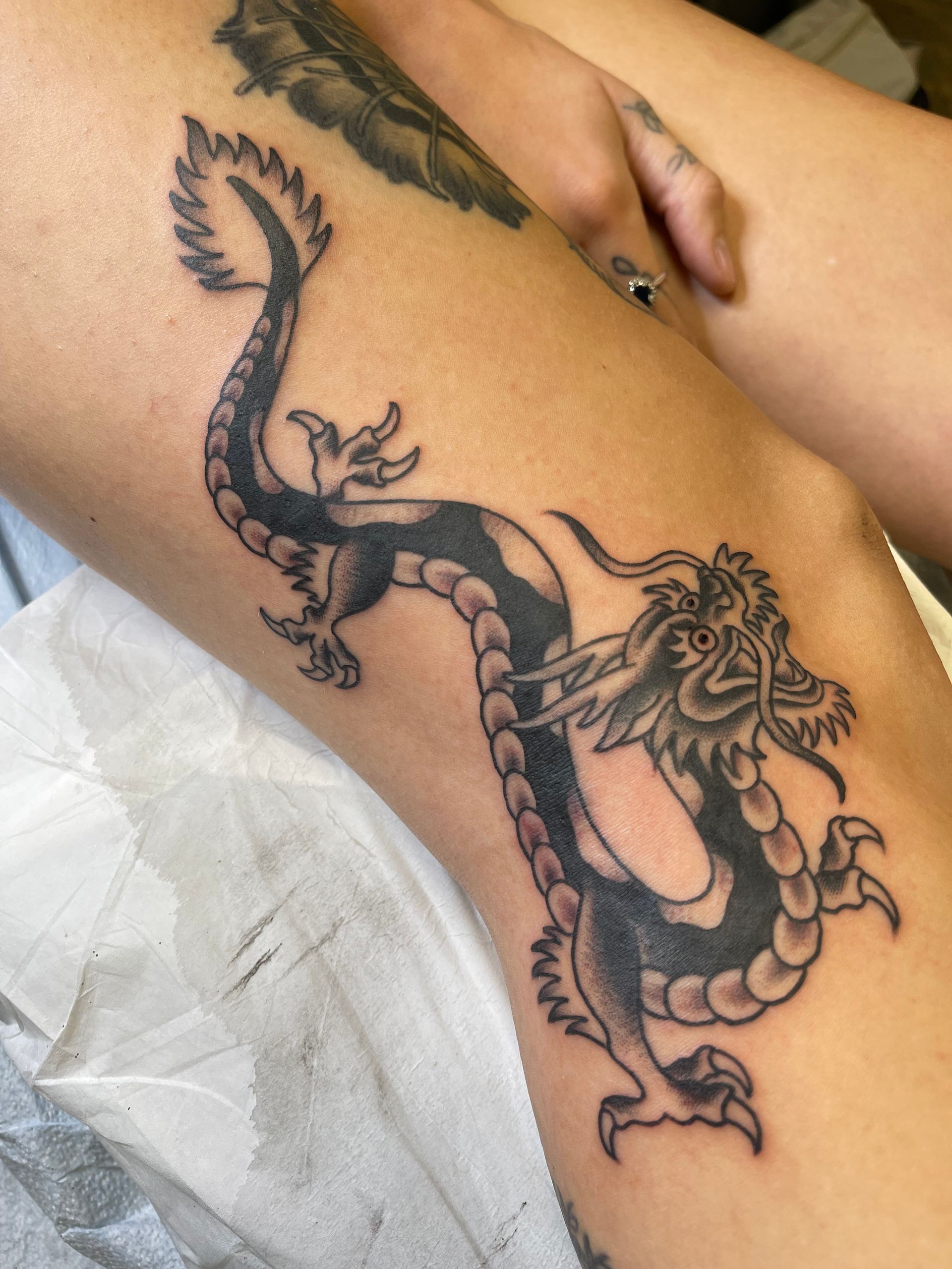 Black dragon tattoo on a person's leg, curled and facing right. The tattoo covers the knee area.