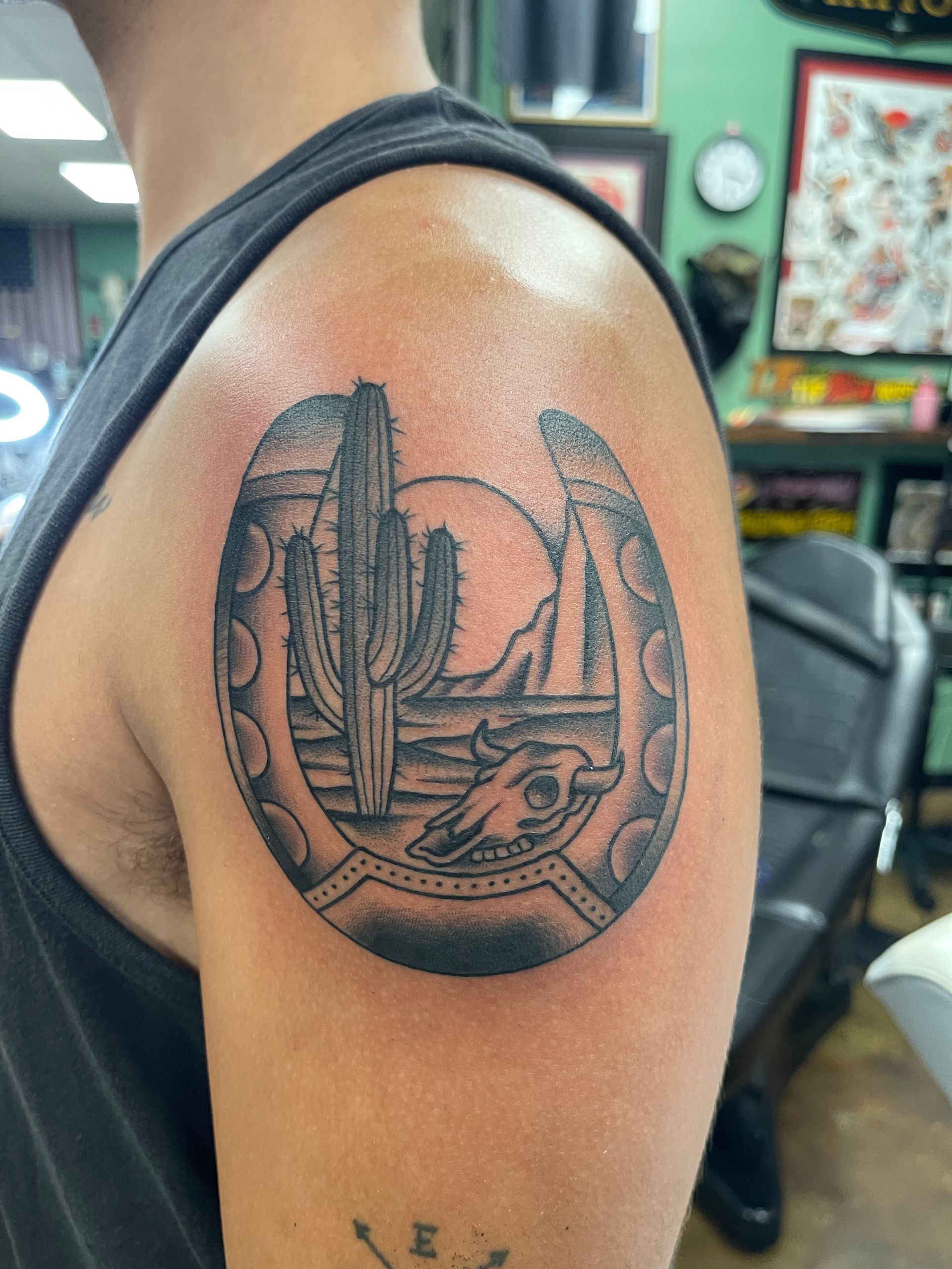 Tattoo of a desert scene inside a horseshoe on a person's upper arm. Features a cactus, skull, mountains.