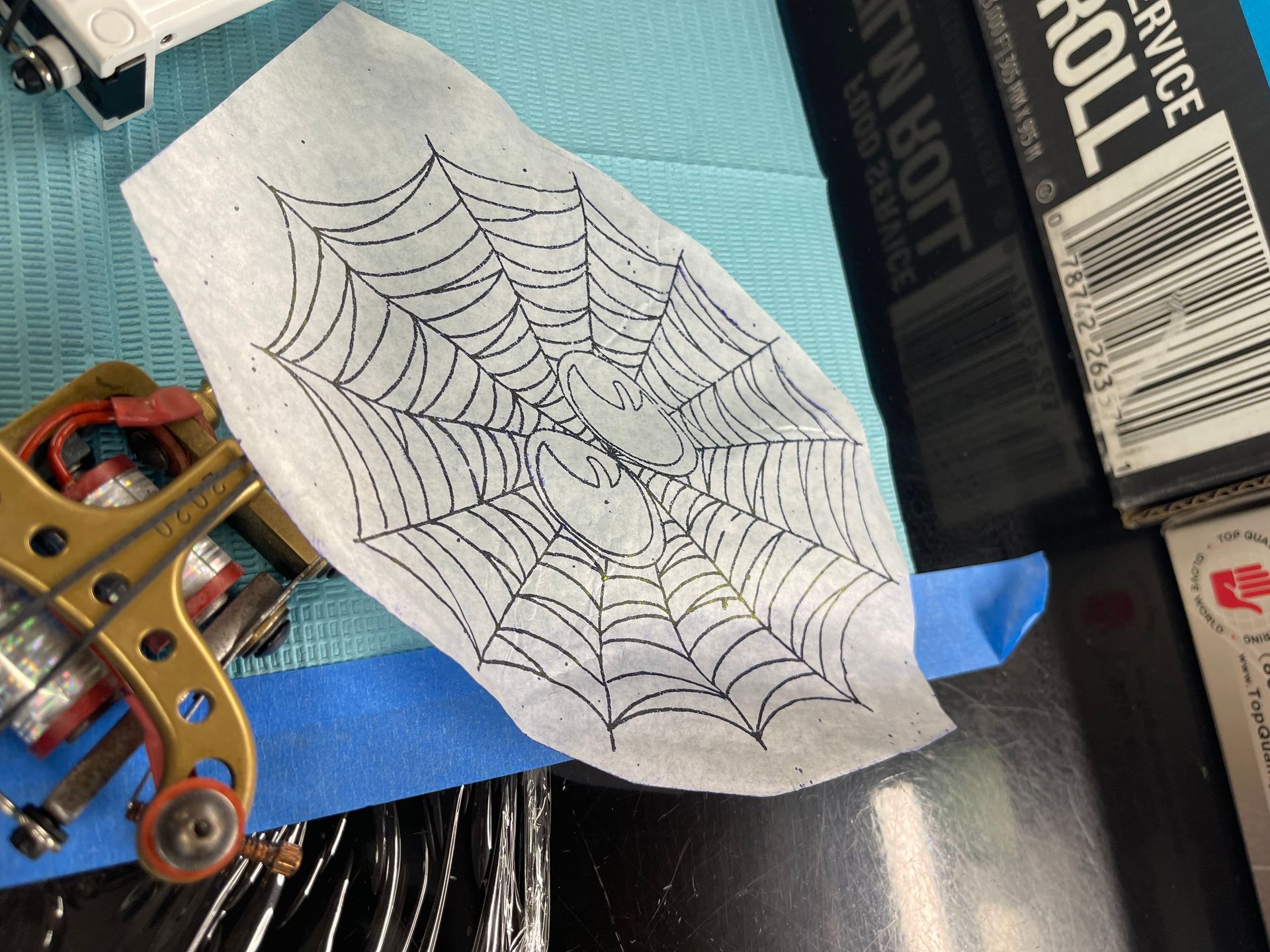 Spiderweb tattoo stencil on a tissue, with a tattoo gun, and supplies.