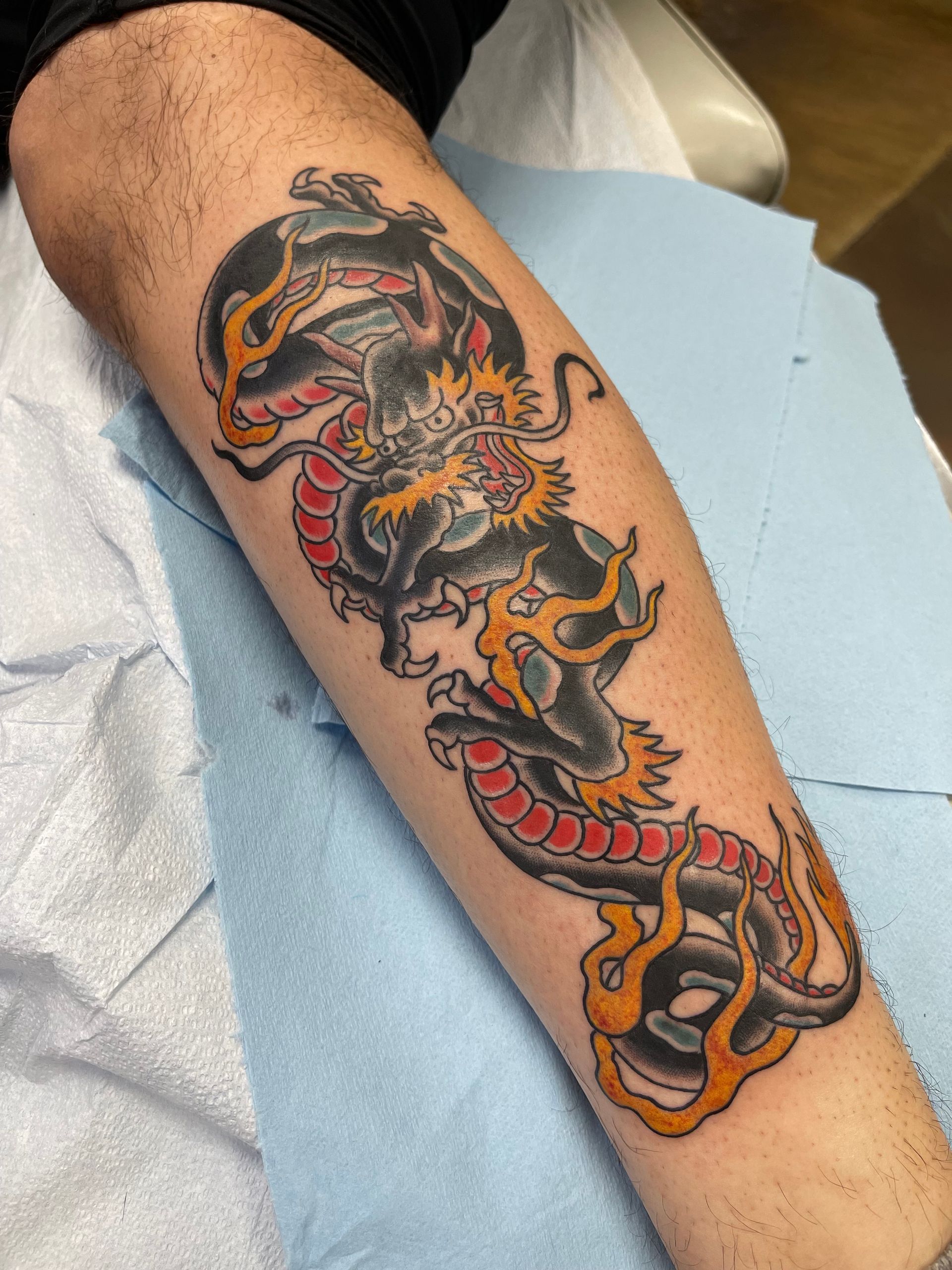 Tattoo of a black dragon with red and orange accents on a person's forearm, lying on a blue surface.