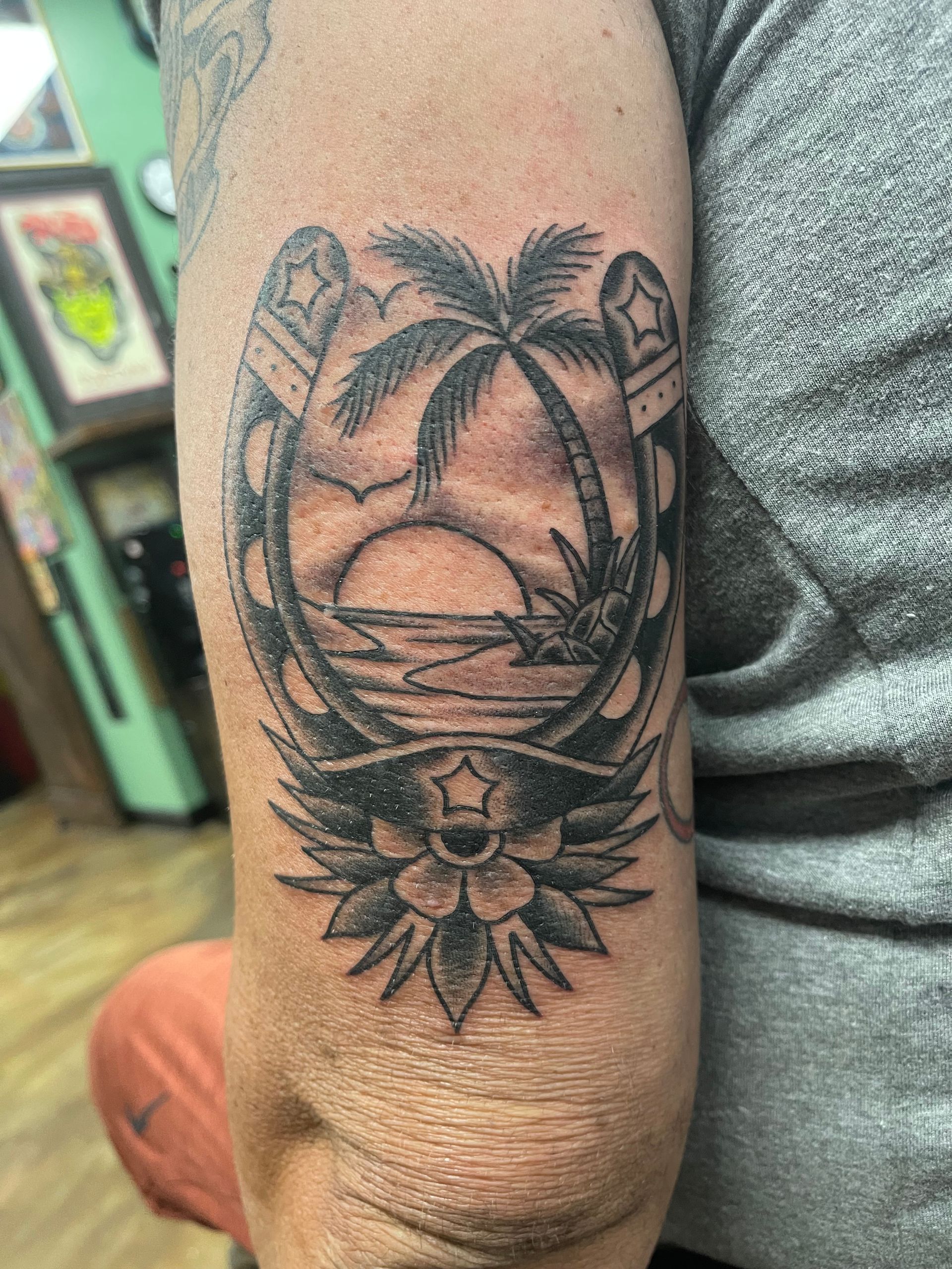 Tattoo on arm depicts a horseshoe with a beach scene: palm tree, sunset, and a flower below.