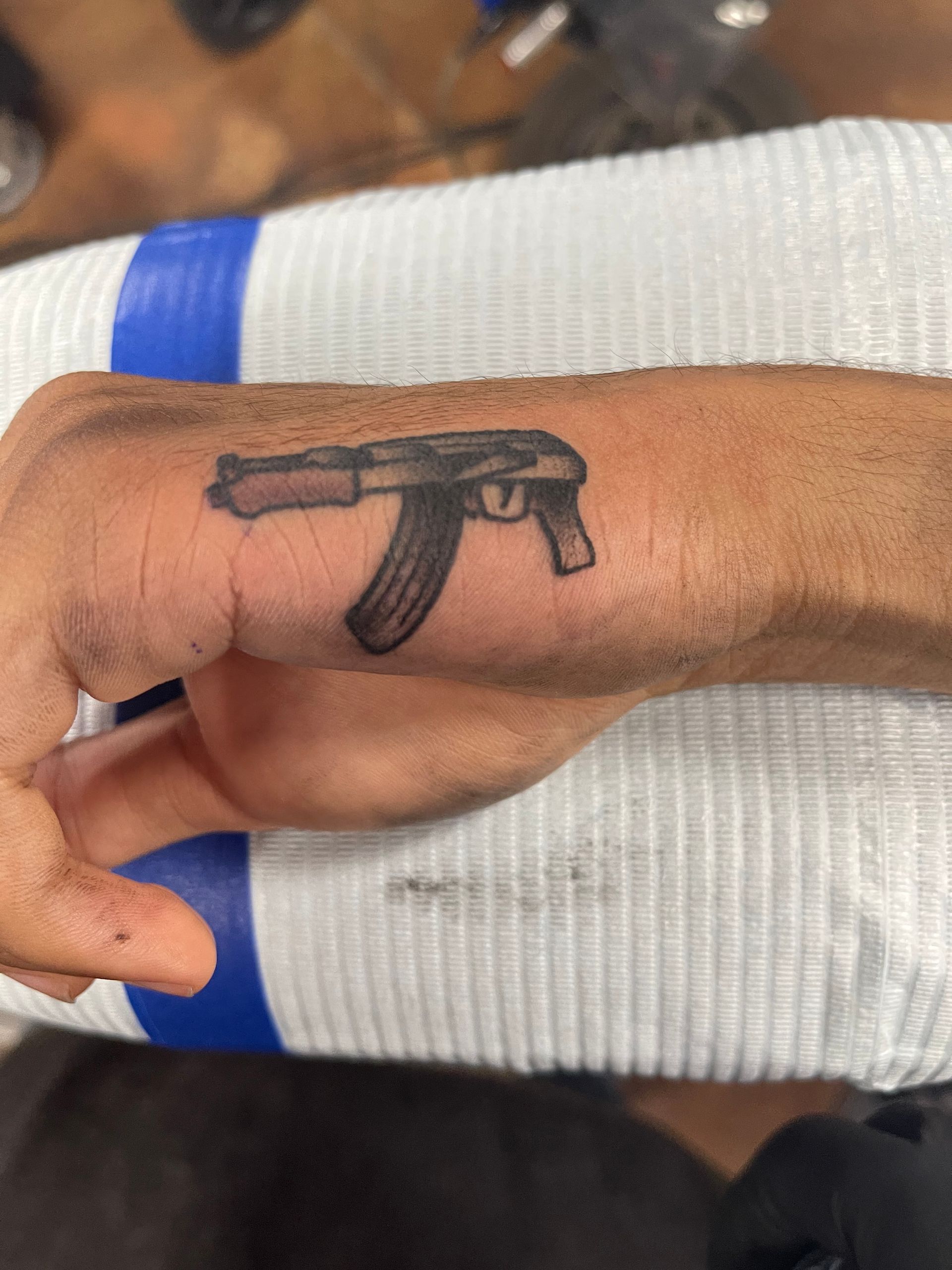 Tattoo of an AK-47 assault rifle on the back of a person's hand. Black ink, visible detail.