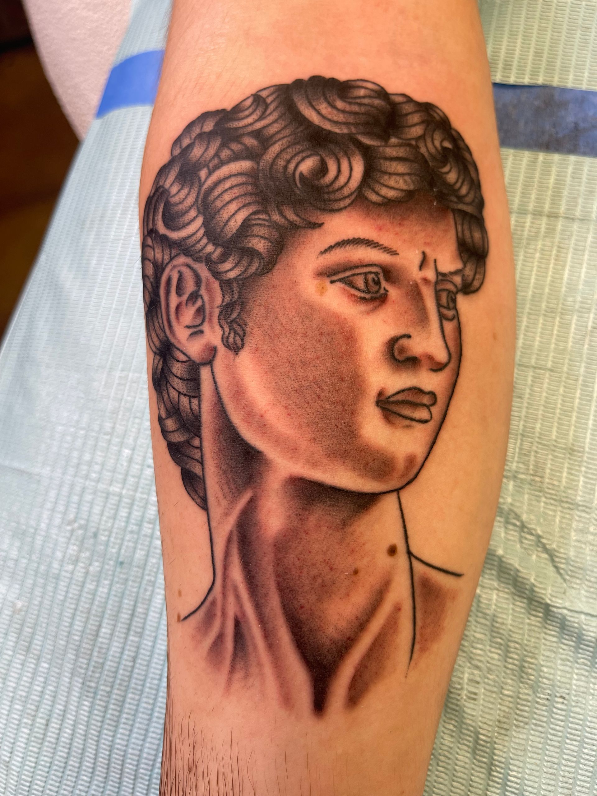 Tattoo of a classical bust on an arm. Black and gray shading depicts the sculpture's curly hair, facial features, and neck.