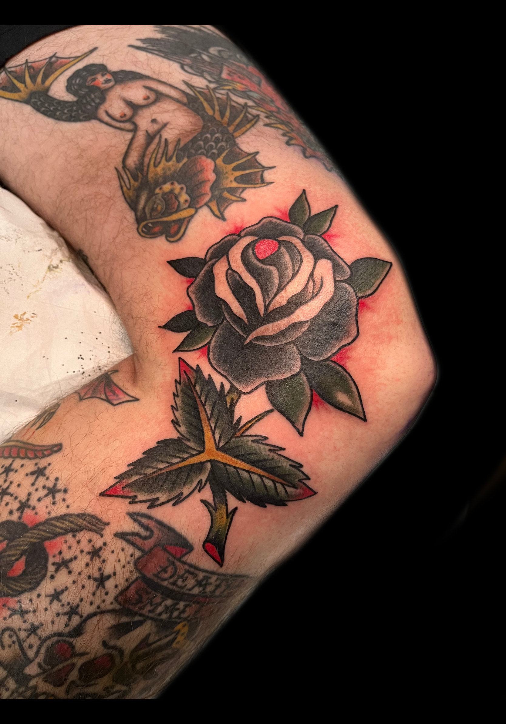 Arm tattoo featuring a black rose, green leaves, and other traditional tattoo designs in red and black.