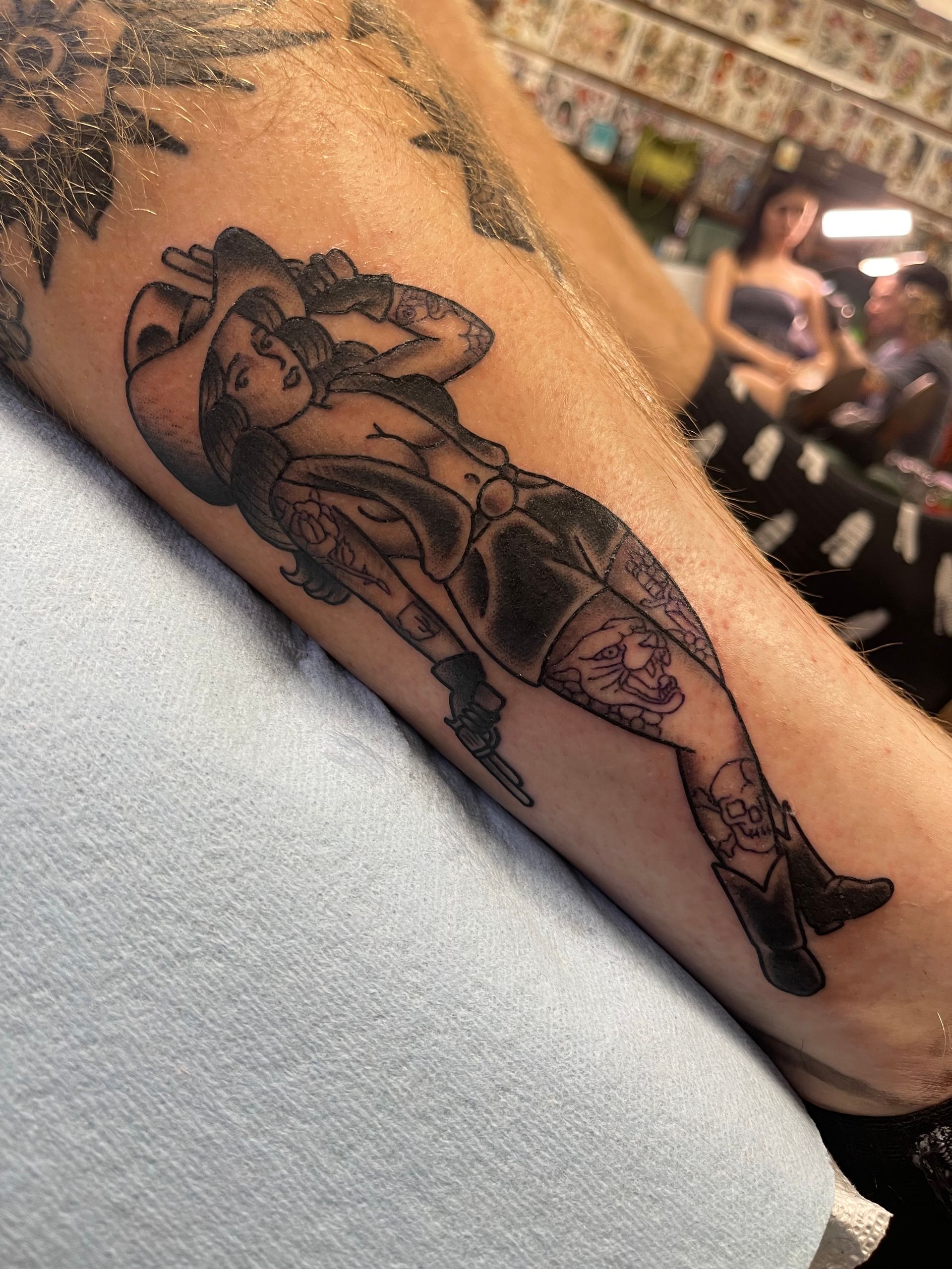 Black and white tattoo of a woman wearing a cowboy hat and boots, lying down on an arm.
