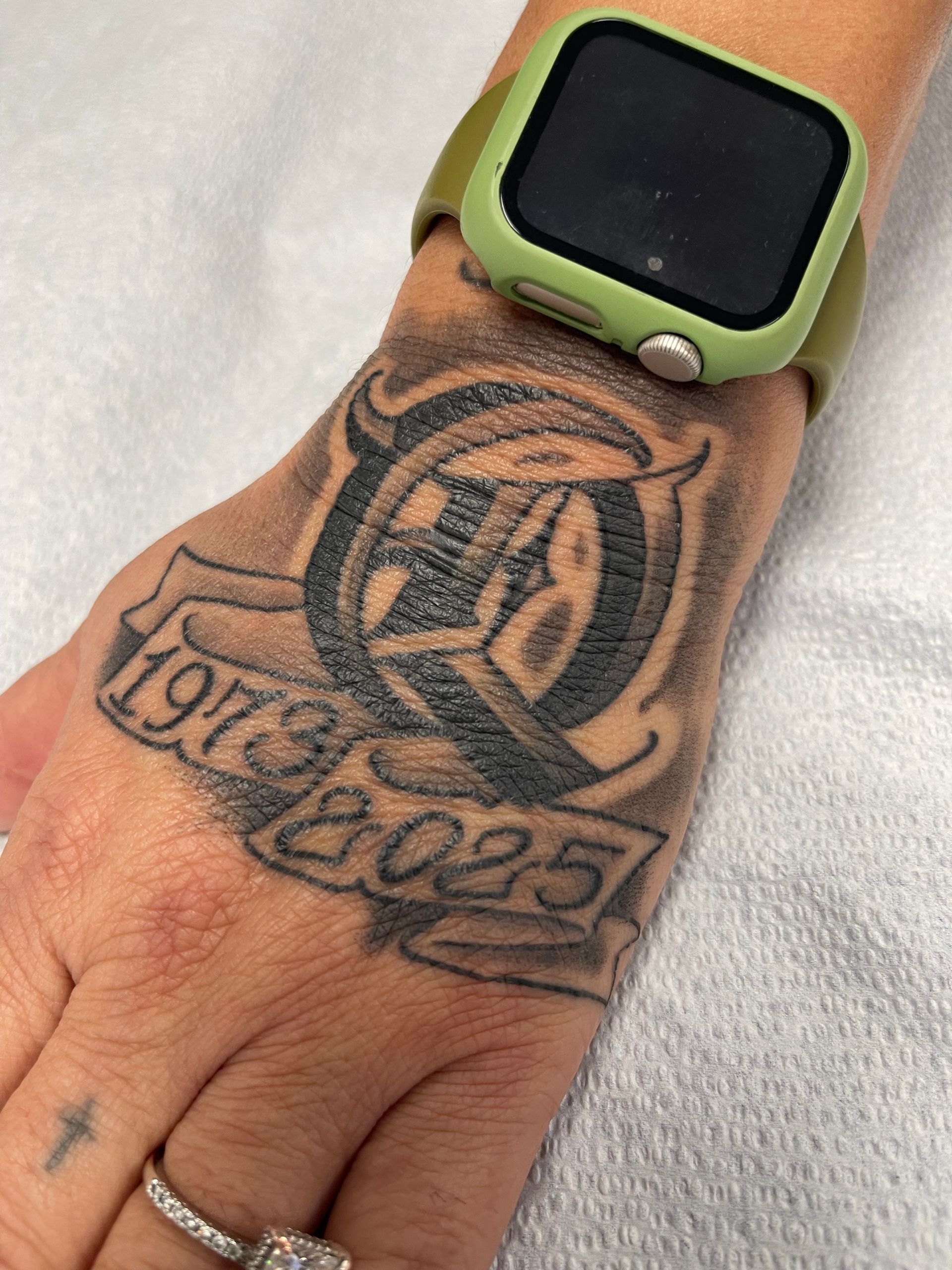 Close-up of a tattooed hand wearing a green smartwatch, resting on a white surface.
