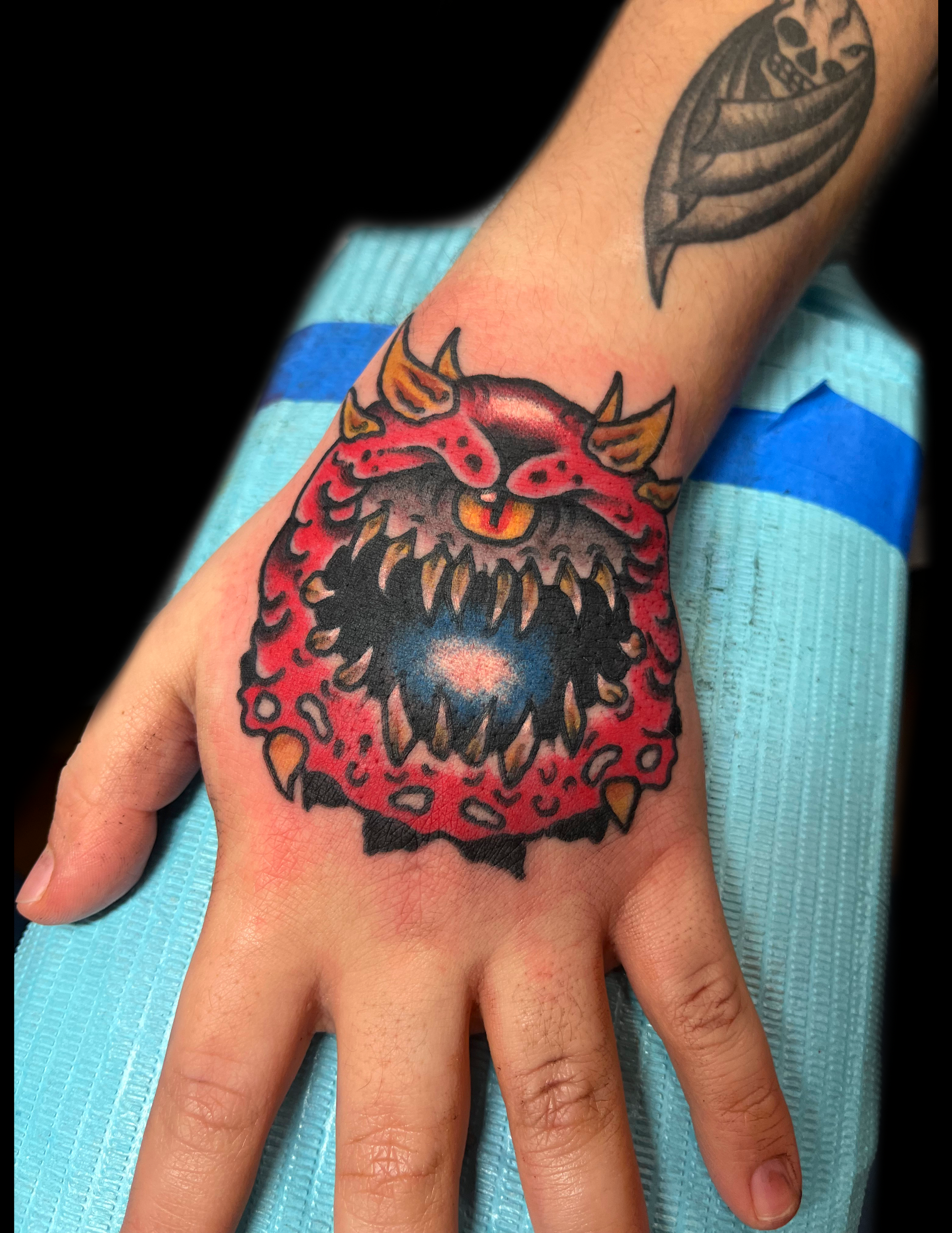 Hand with a red, monster-like tattoo featuring sharp teeth and horns. A smaller tattoo is visible on the arm.