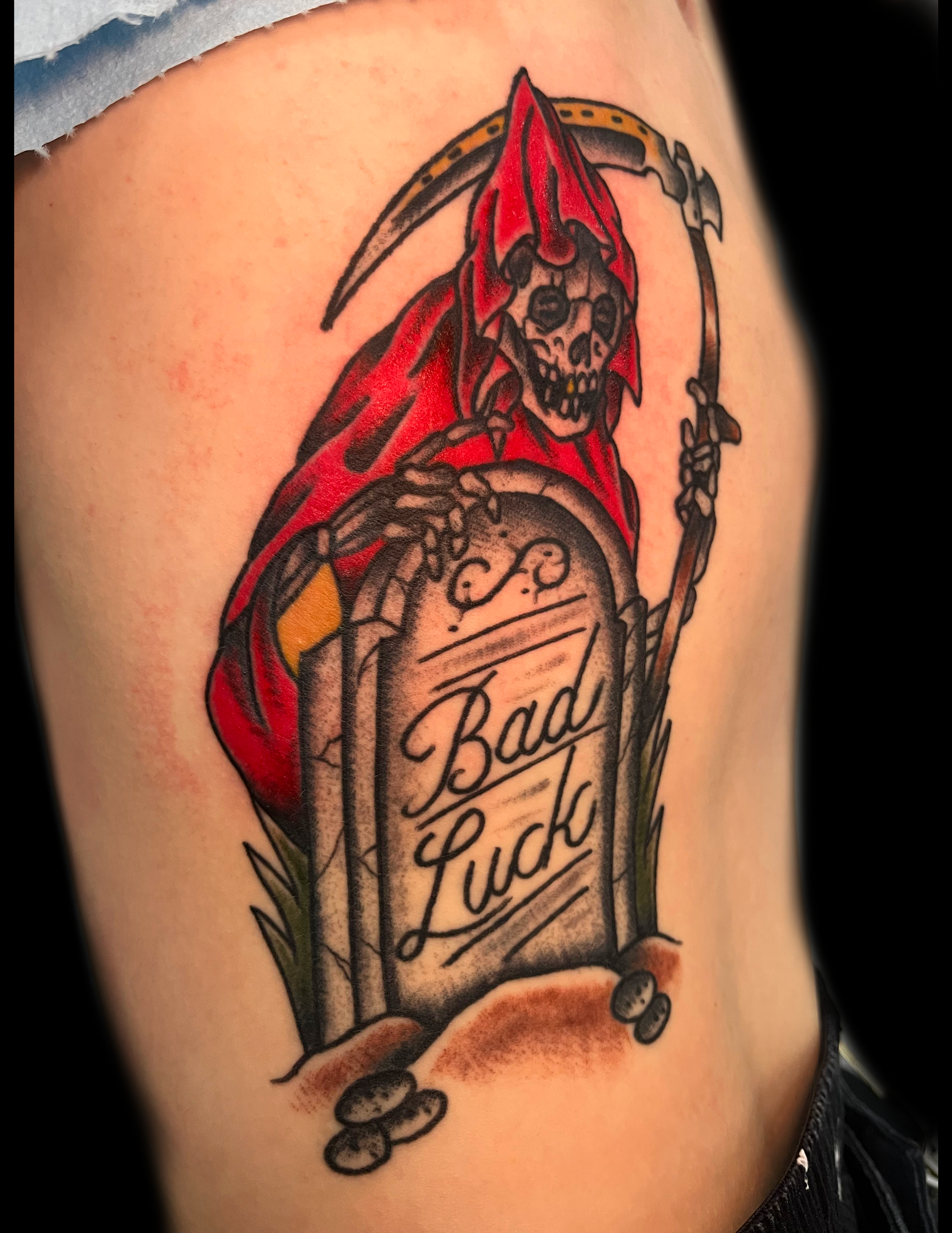 Grim reaper in red cloak with scythe, standing by tombstone that says 