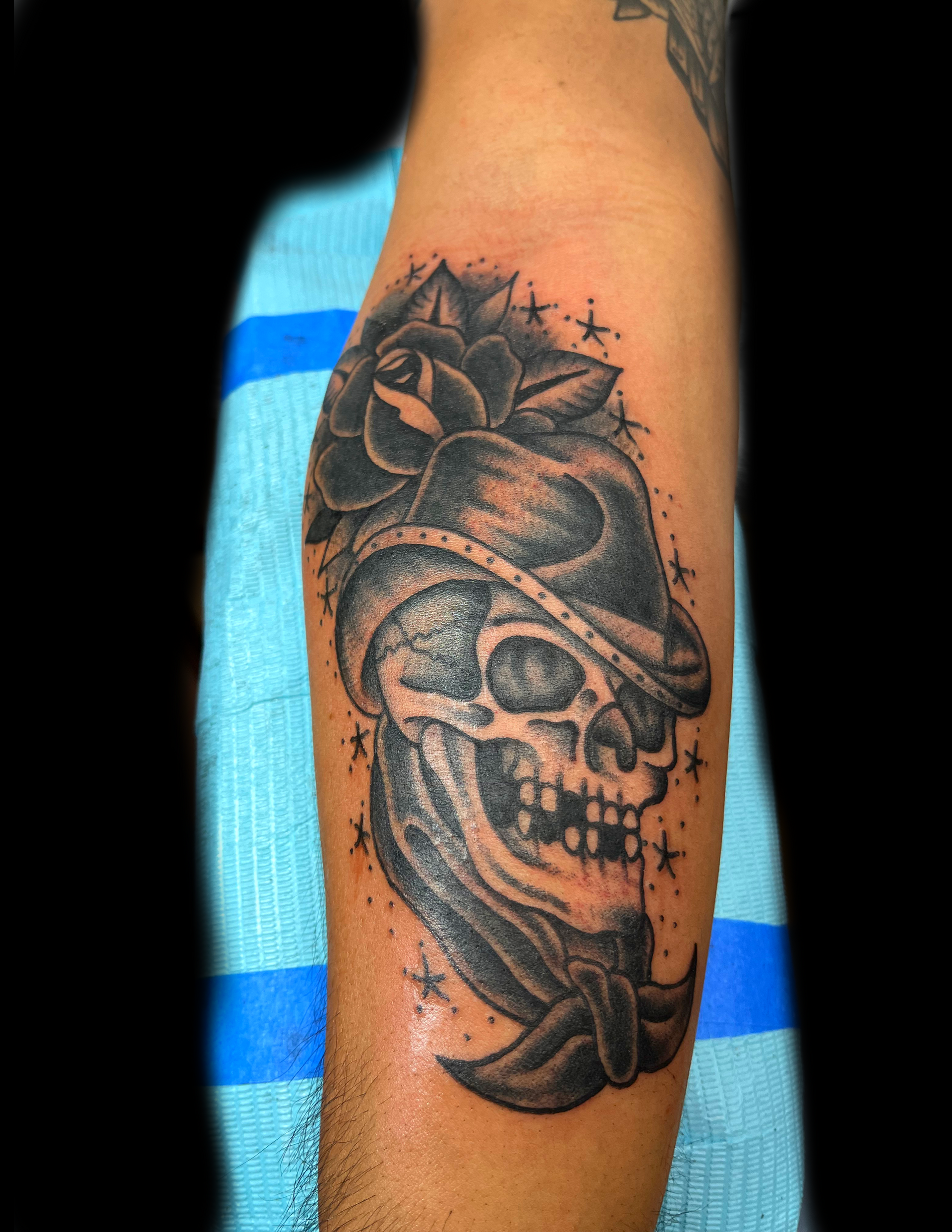 Tattoo of a skull wearing a hat and bandana, with a rose above the hat, done in black ink on an arm.
