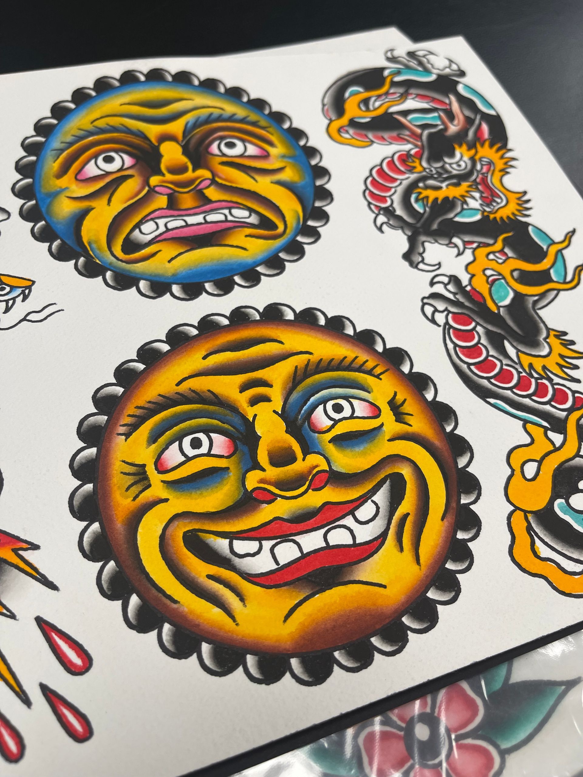 Tattoo flash sheet with two smiling sun faces, a dragon, and red teardrops, vibrant colors.