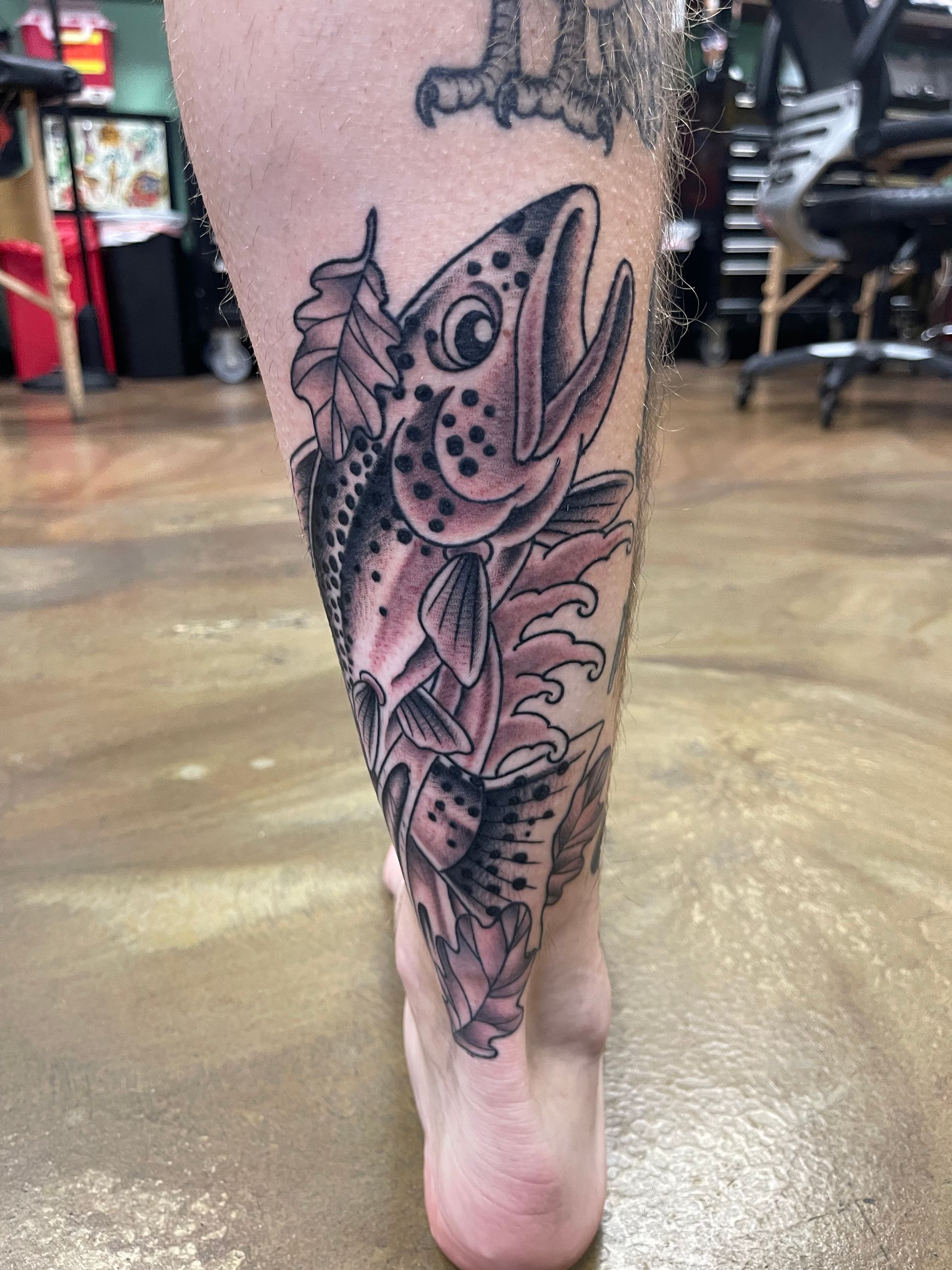 Tattoo of a salmon with open mouth and spots on a leg, surrounded by leaves and water swirls.