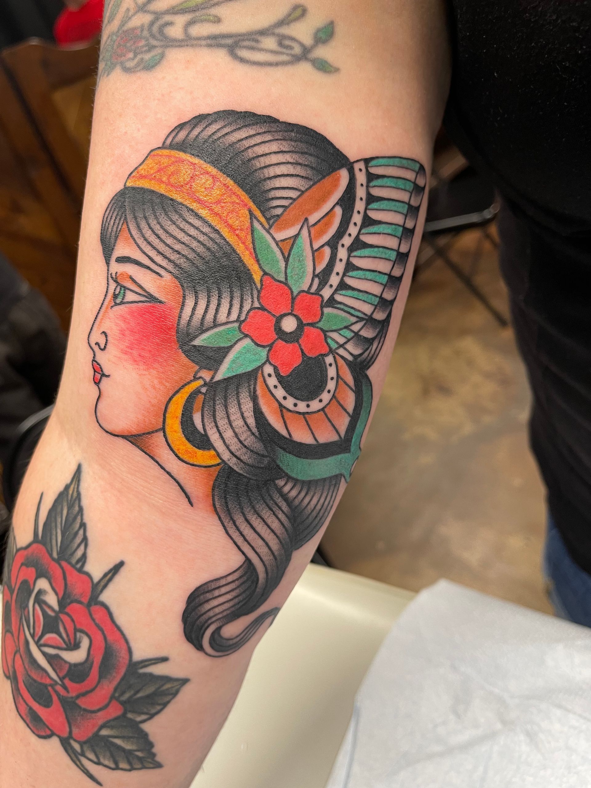 Tattoo of a woman in profile, wearing an orange headband and flower, on an arm. A red rose tattoo is below it.