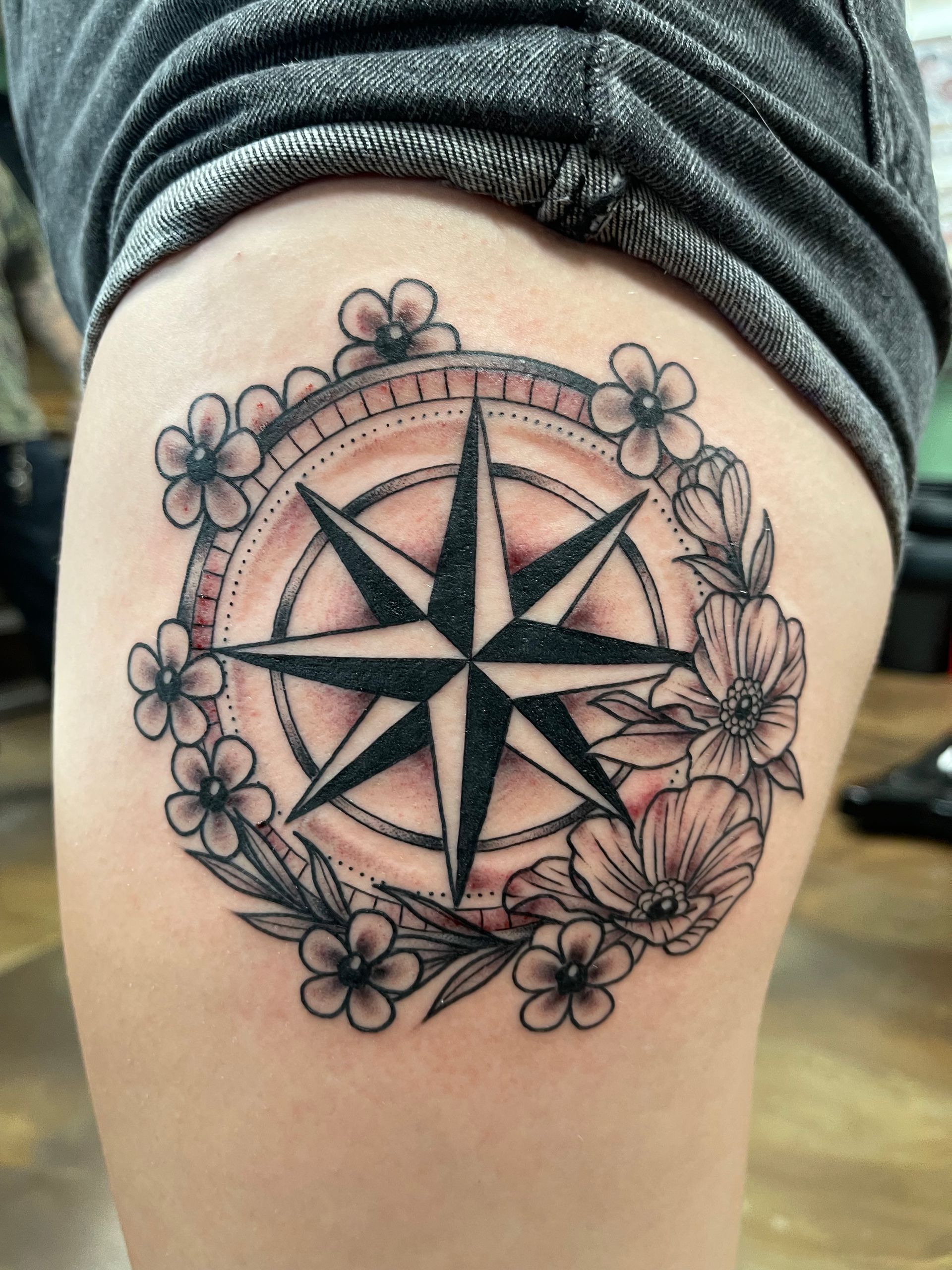 Tattoo of a black compass rose surrounded by flowers on a person's thigh.