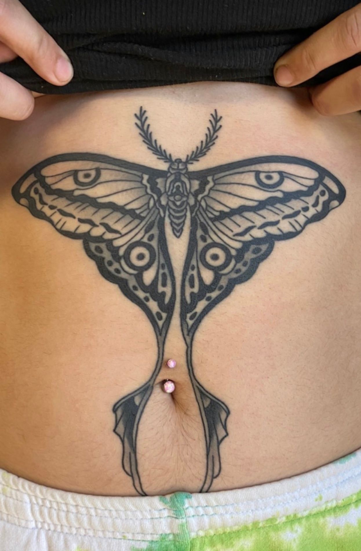 Tattoo of a moth on a person's abdomen, with a navel piercing.