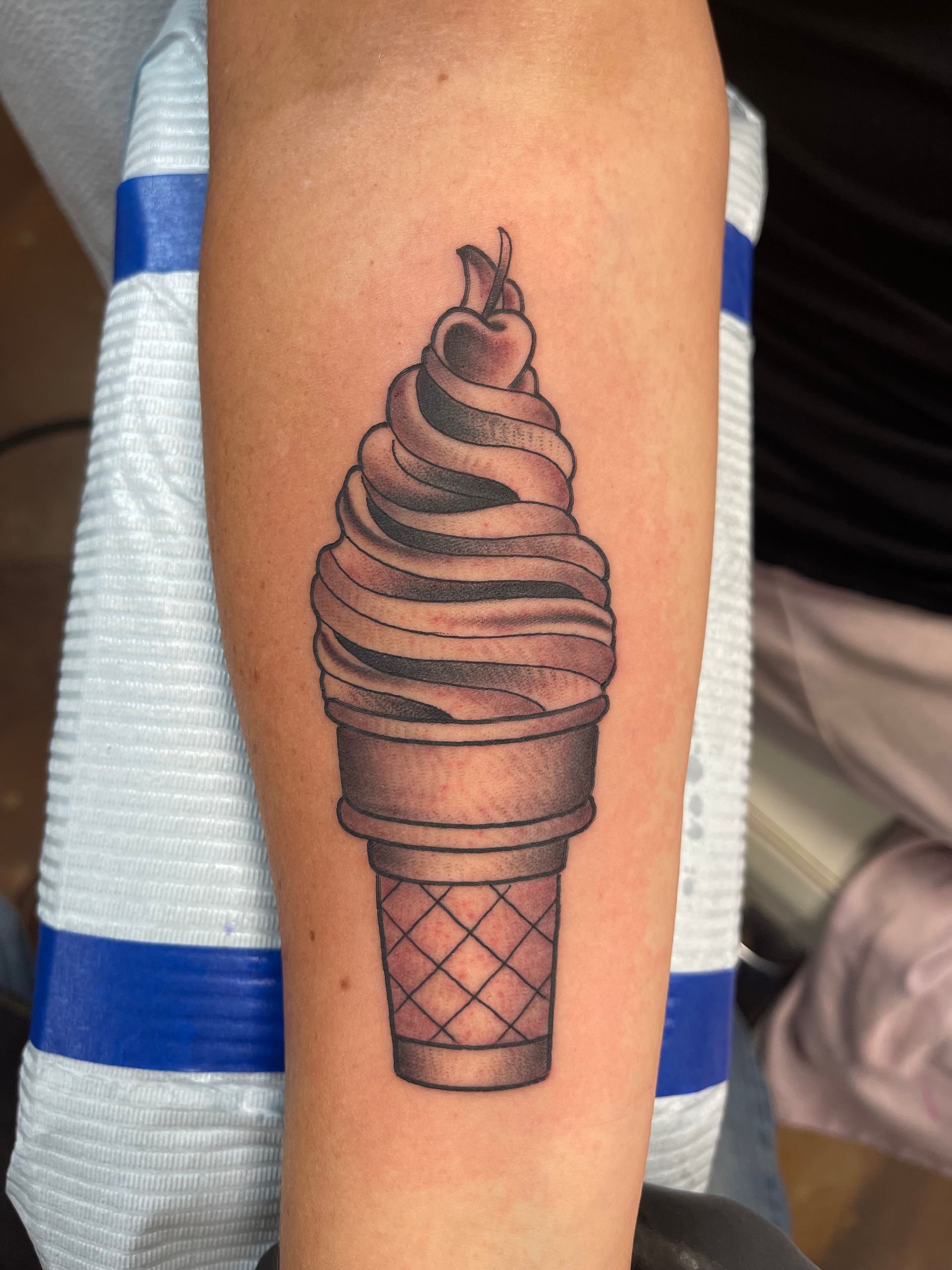 Tattoo of a soft-serve ice cream cone with a cherry on top; on a forearm.