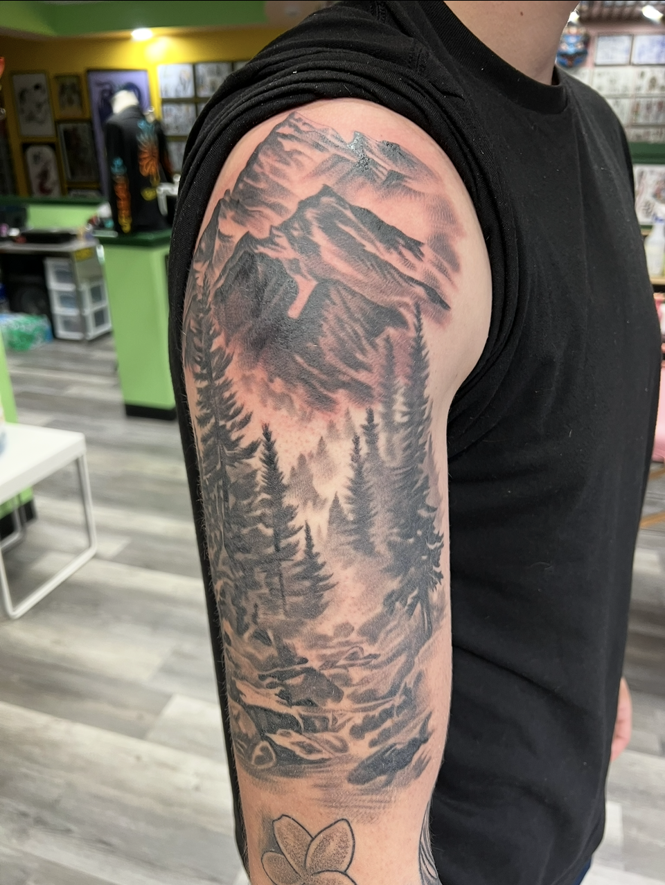 Arm tattoo of a pink mountain and pine forest on a person in a black sleeveless shirt