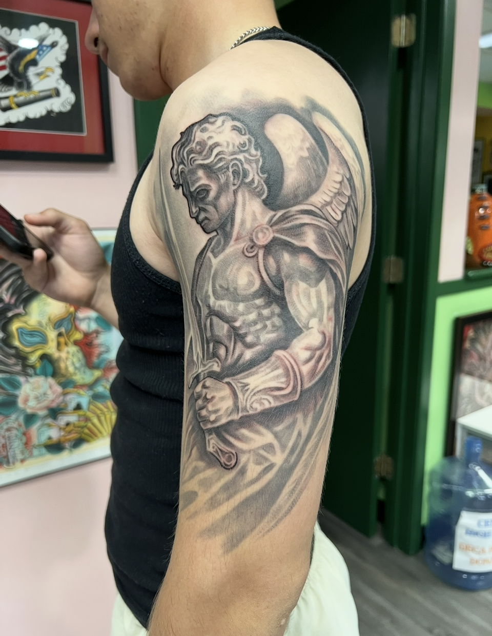 Person showing a detailed black-and-gray angel tattoo on the upper arm in a tattoo studio