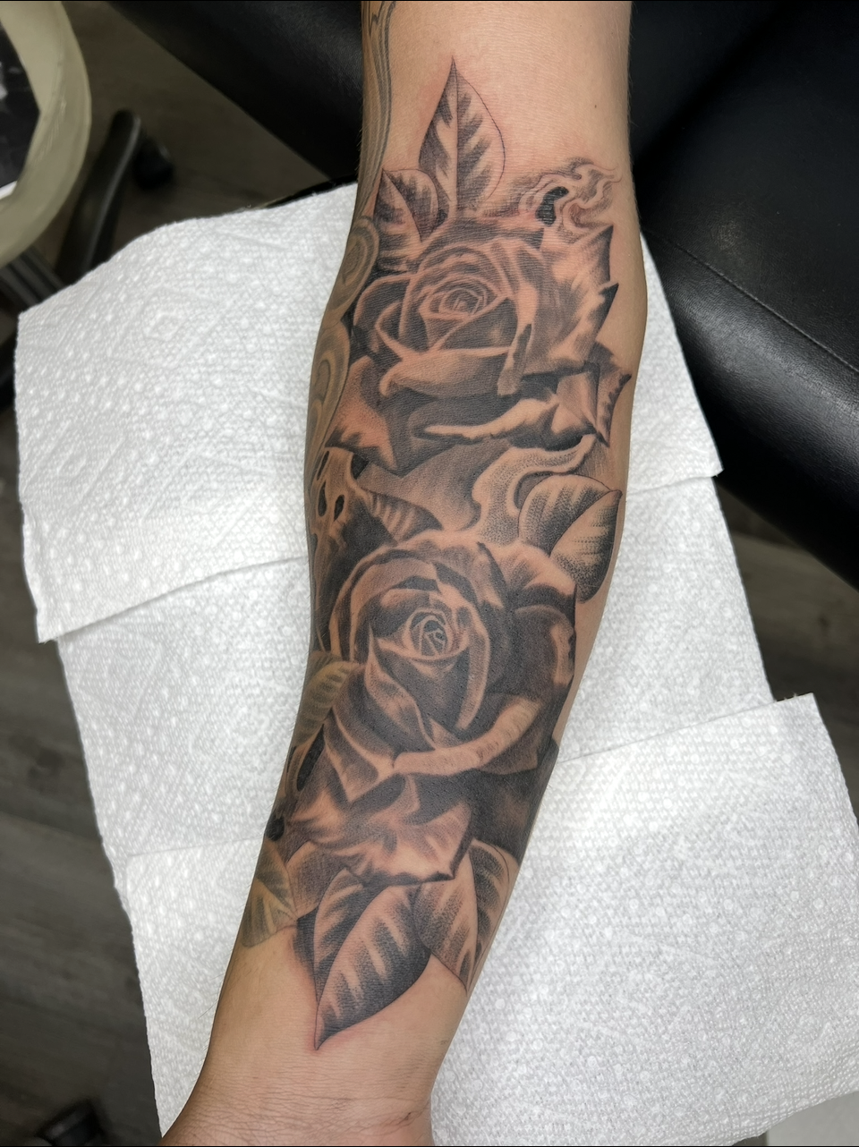 Forearm with a black and gray rose tattoo sleeve on white paper towel and a black chair in the background