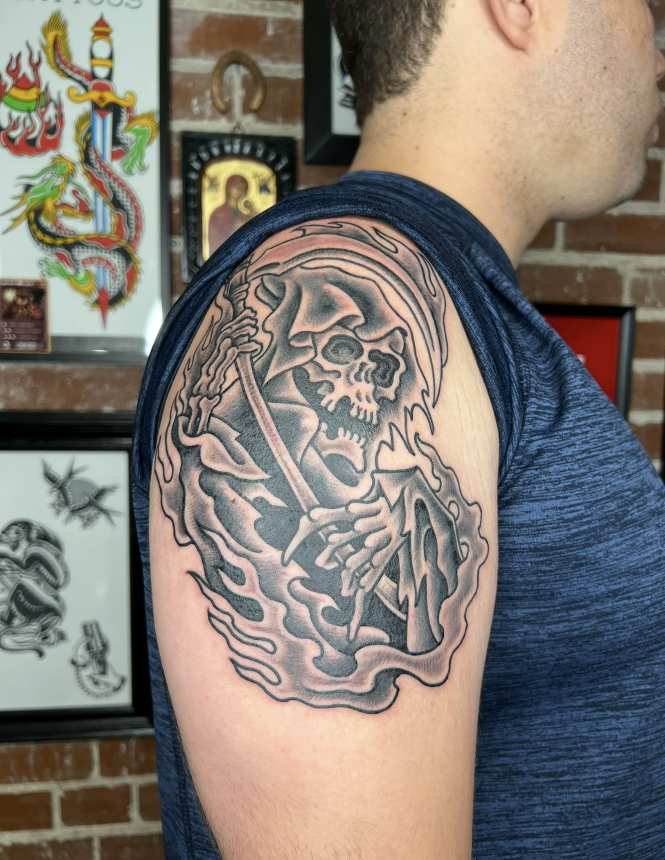 Arm tattoo of a grim reaper on a person’s upper arm against a brick wall and framed artwork