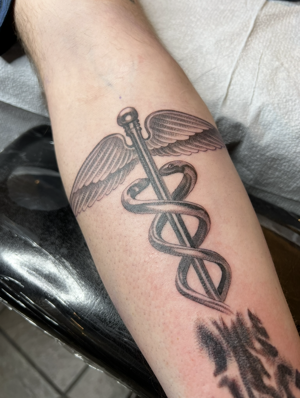 Forearm tattoo of a caduceus with wings in black ink, shown close up on skin
