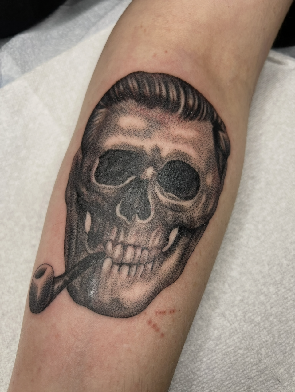 Black-and-gray skull tattoo on a forearm with dark hair combed back and a small smoking pipe